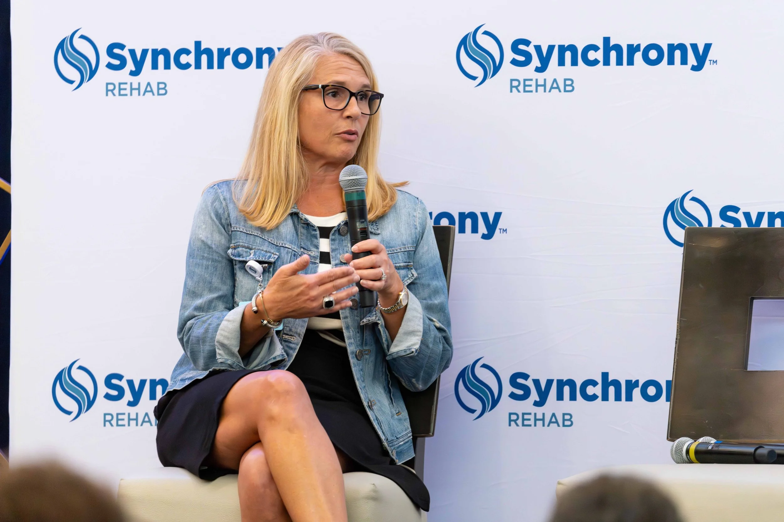 A woman with blonde hair, wearing glasses, a denim jacket, and a black-striped shirt, is sitting and speaking into a microphone at a panel event. The background displays the logo and text for Synchrony Rehab.
