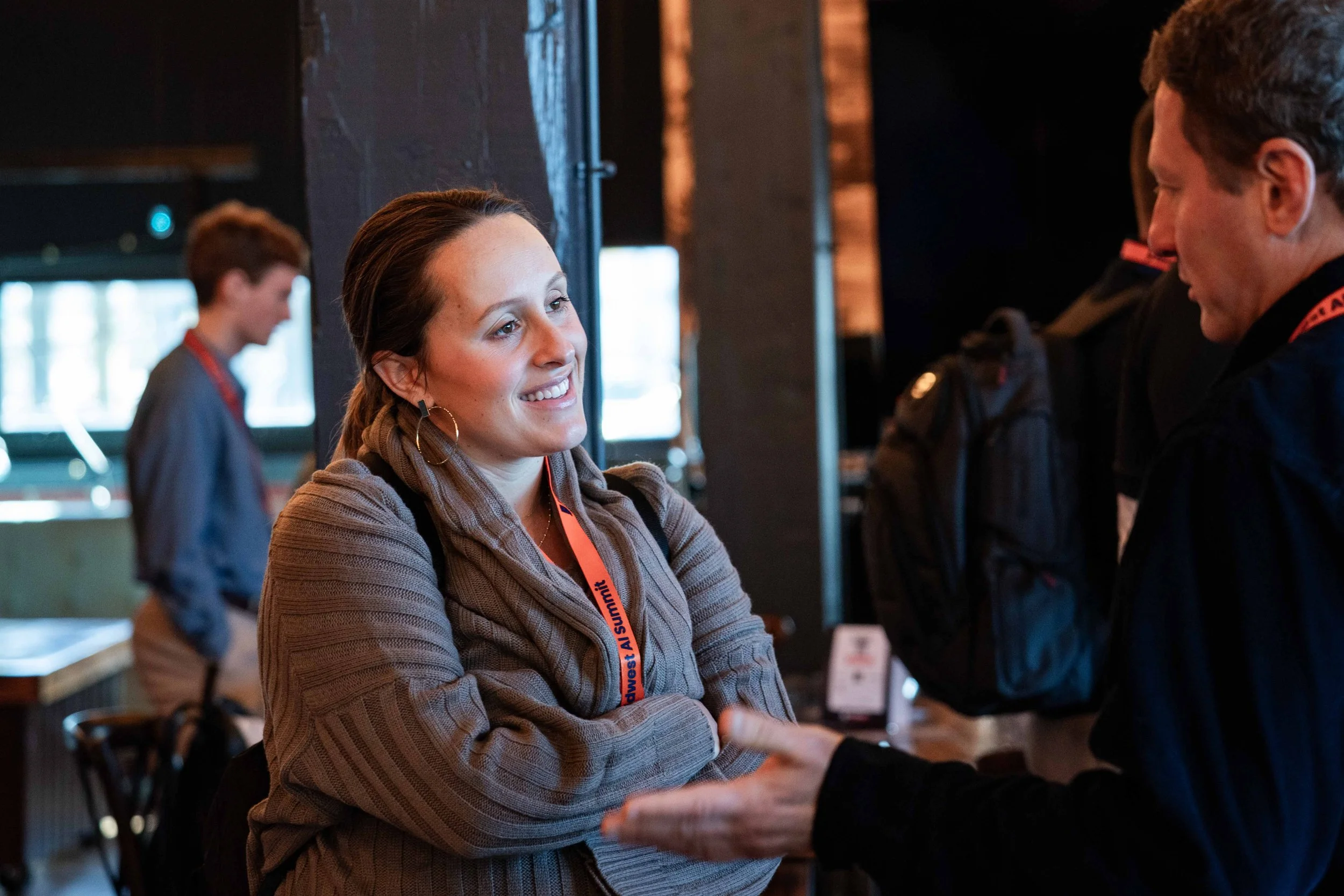 A woman smiling and talking to a man at an indoor event, with a woman in the background.