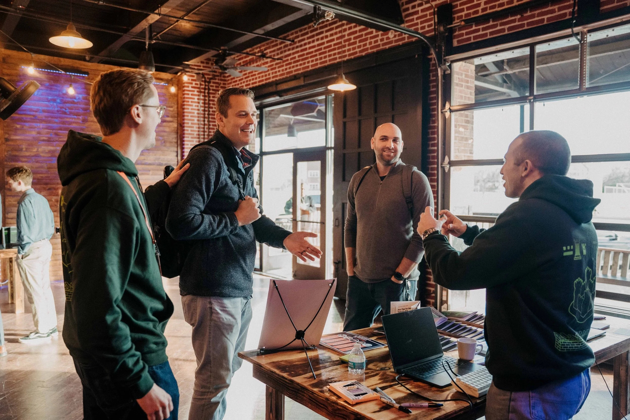 Four men engaging in conversation inside a rustic, modern cafe with brick walls and large windows, one man smiling and gesturing while another listens attentively near a table with laptops and pamphlets.