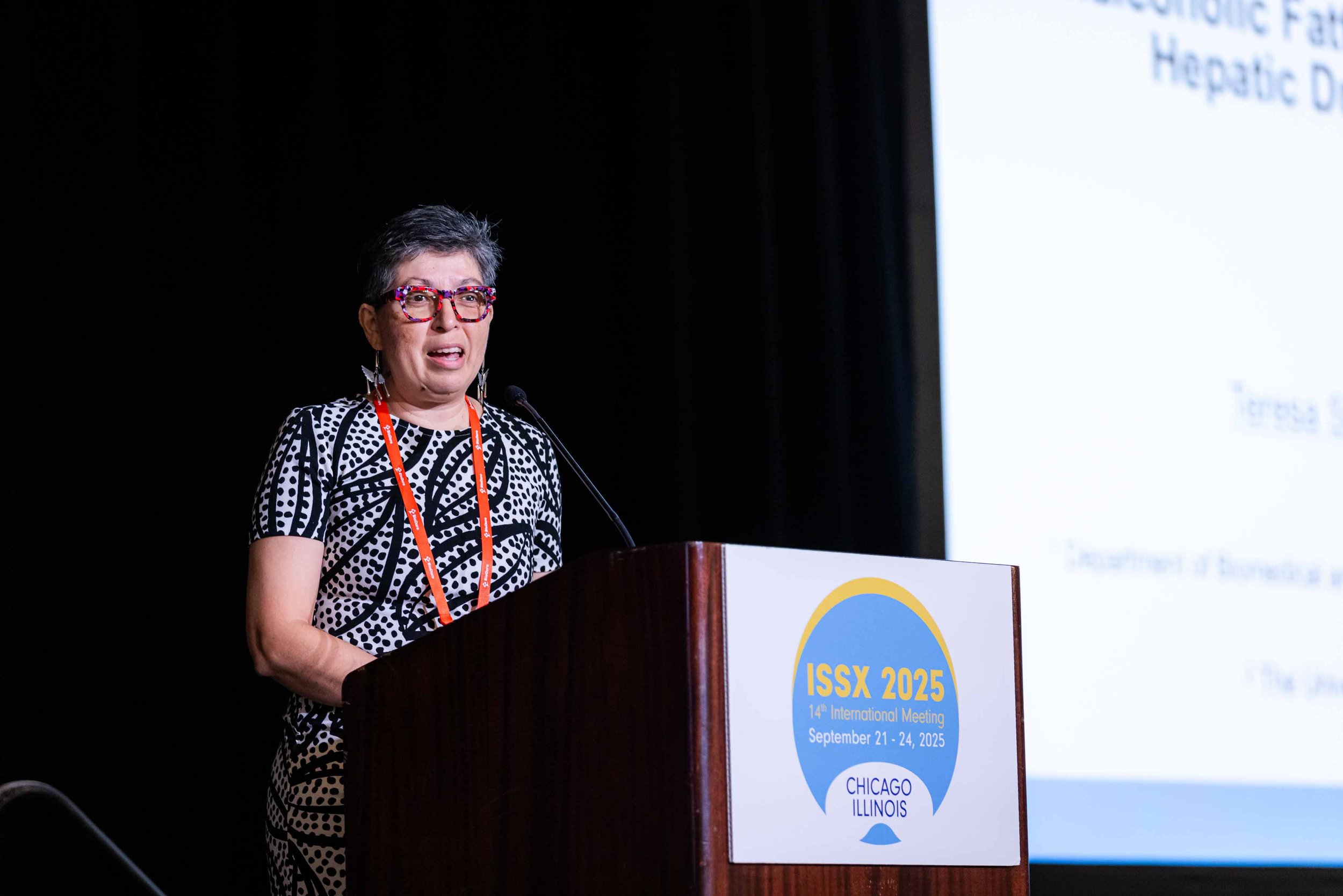 A woman with short dark hair, glasses with a red and black pattern, wearing a black and white patterned dress, speaking at a podium during the ISSX 2025 conference in Chicago, Illinois, with a presentation slide visible in the background.