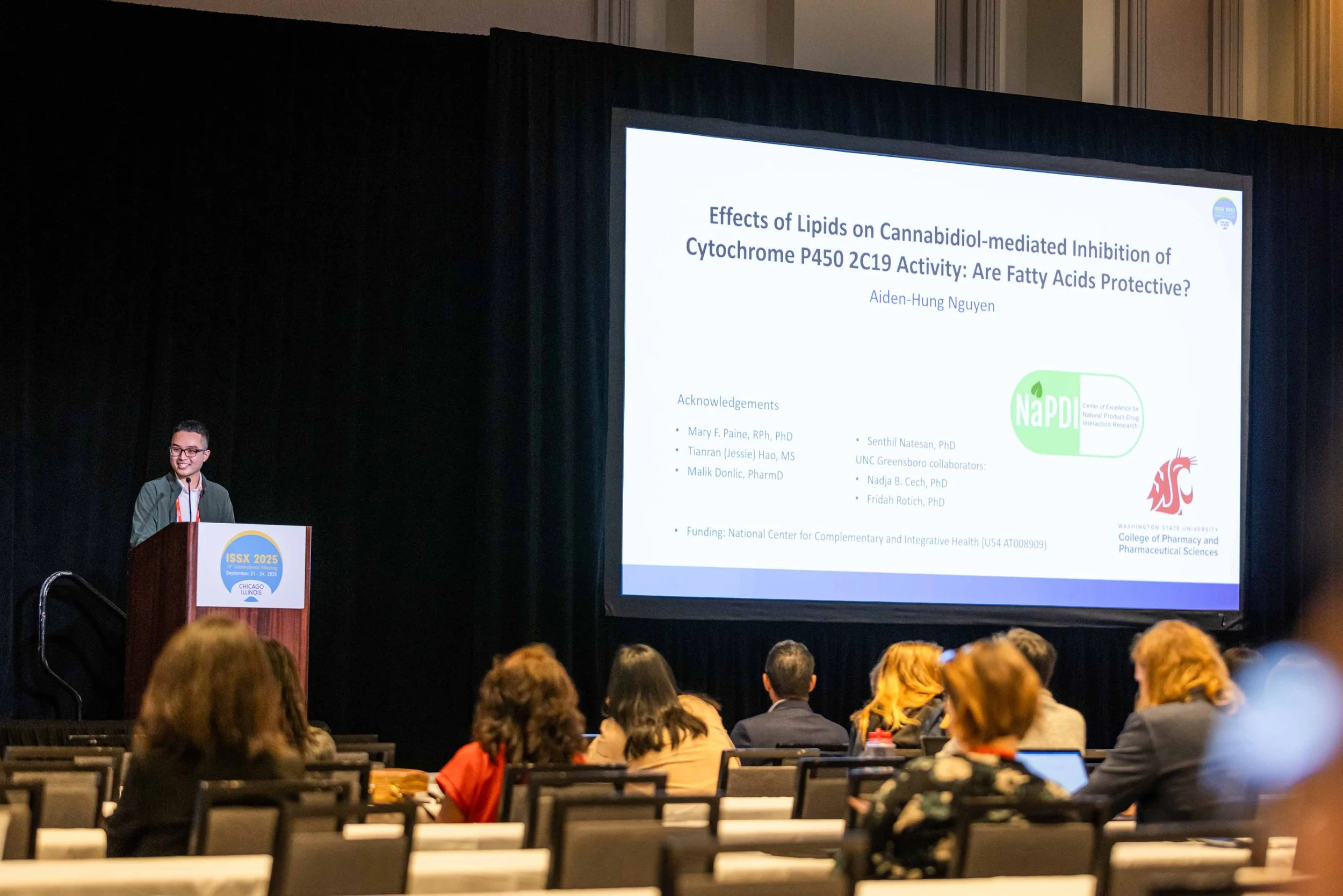 Young male presenter standing at a podium with a microphone, giving a presentation at a conference called ISSX 2025, with a large screen displaying a scientific research slide about effects of lipids on cannabinoids, in a conference hall filled with 