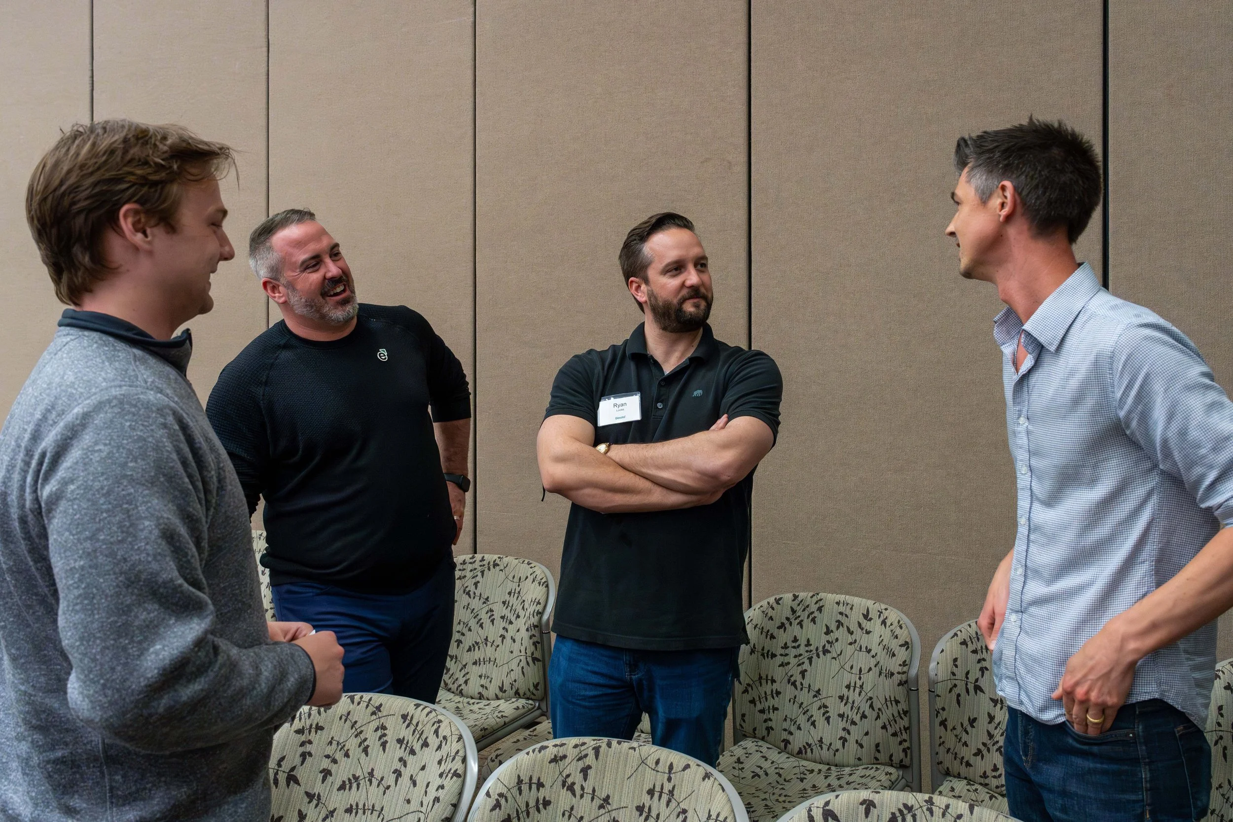 Four men engaged in conversation in a conference room, with chairs and a beige wall in the background.