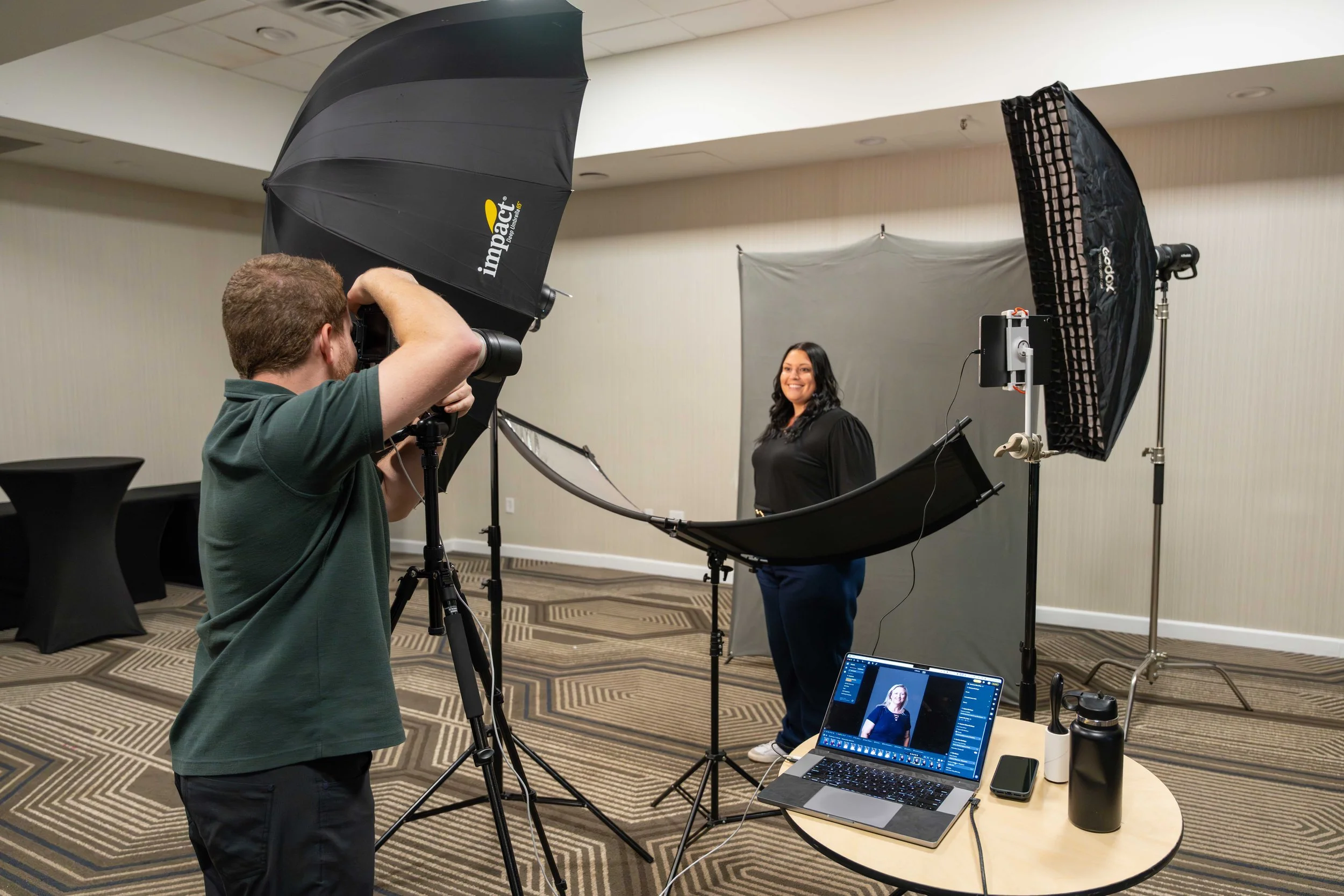 A woman with long dark hair stands in a photography studio, smiling at the camera. A photographer is taking her picture with professional lighting equipment and a backdrop. A laptop on a table displays a live preview of the photo shoot.