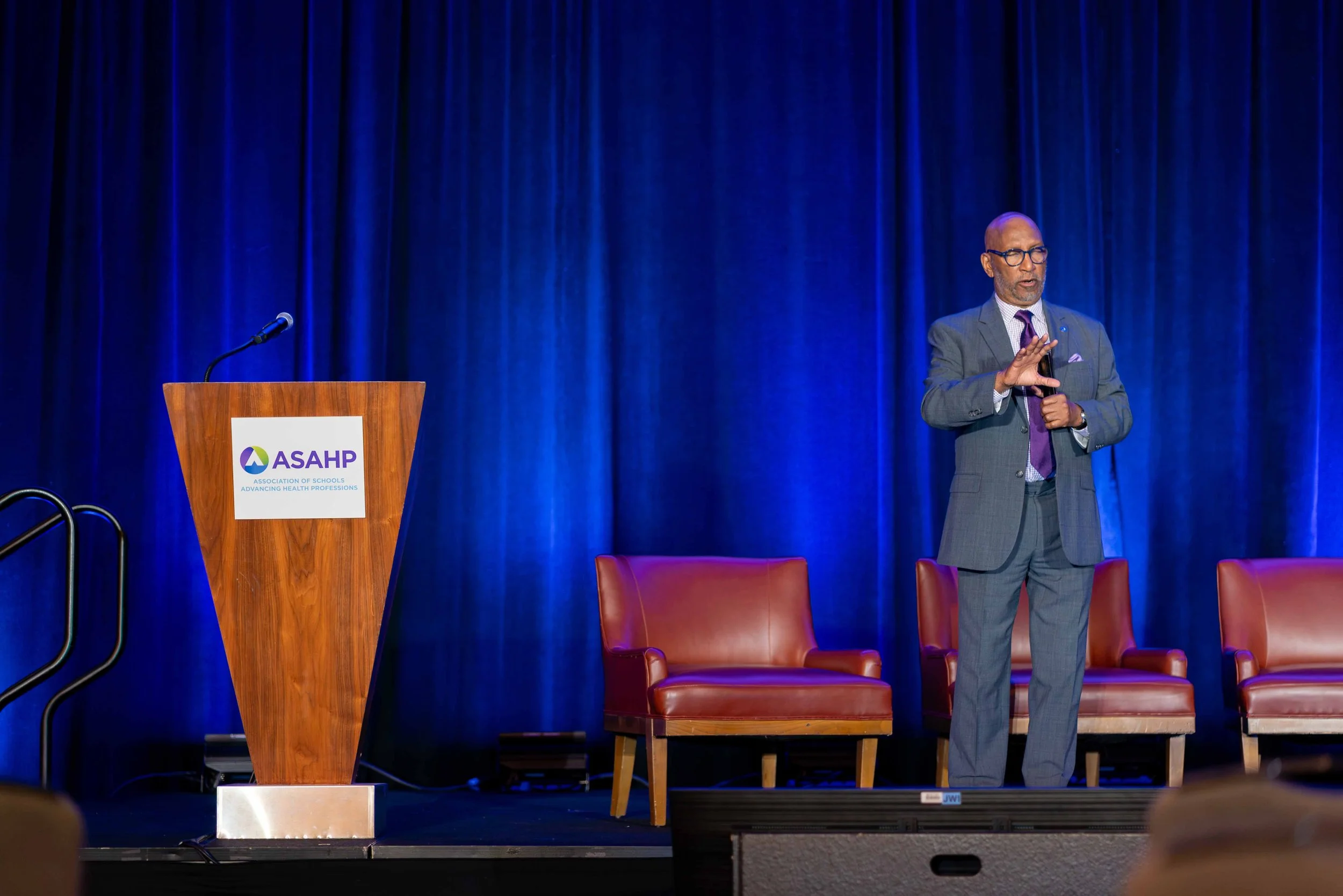 A man in a gray suit giving a presentation on a stage with a blue curtain backdrop. Next to him is a wooden podium with an ASAPH logo, and there are red chairs behind him.