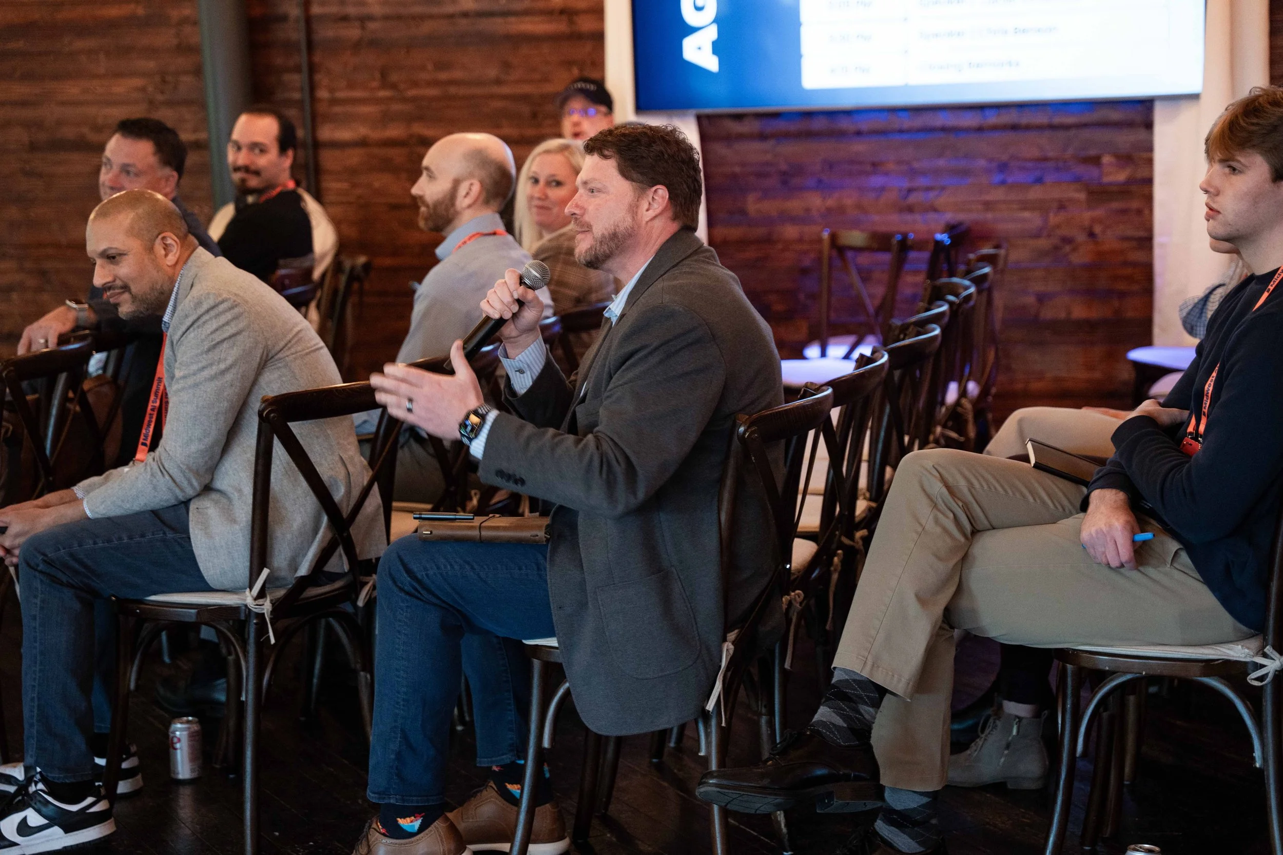 Audience members at a conference, with one man speaking into a microphone, seated in a room with wooden walls and a large screen behind them.