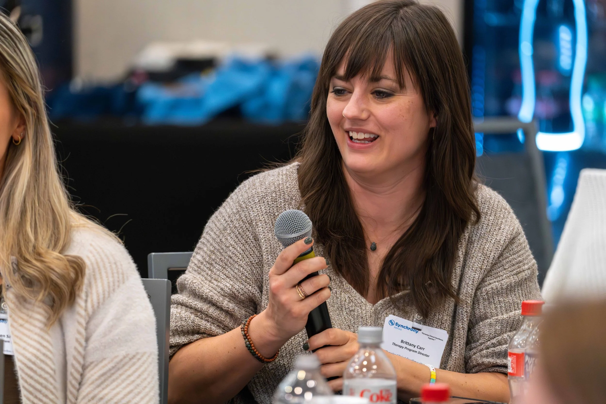 A woman with brown hair, wearing a beige sweater, holding a microphone, and sitting at a conference table with water bottles and a name tag.