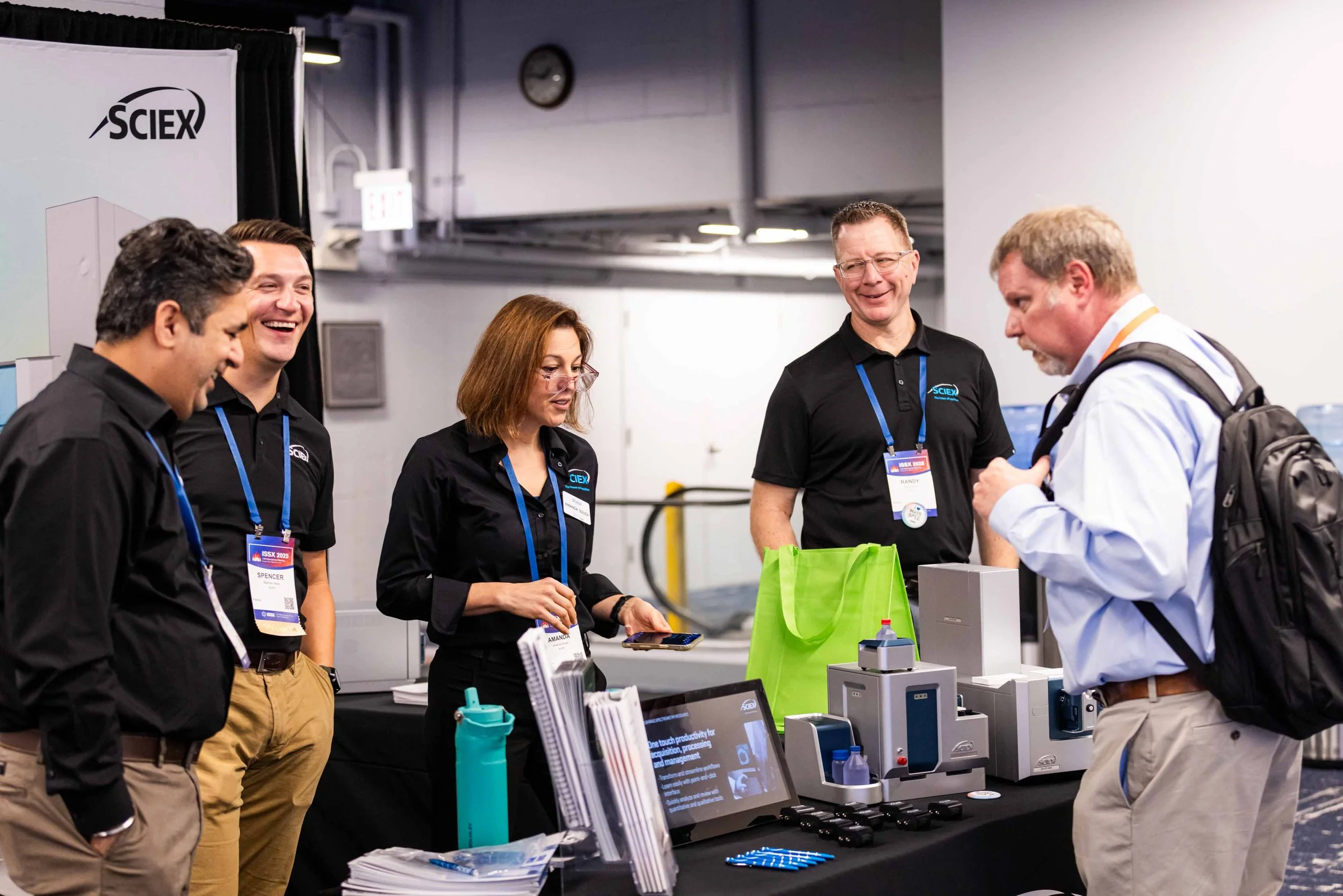 Group of five people at a booth during a conference, engaging with a man with a backpack, with display items and promotional materials on the table.