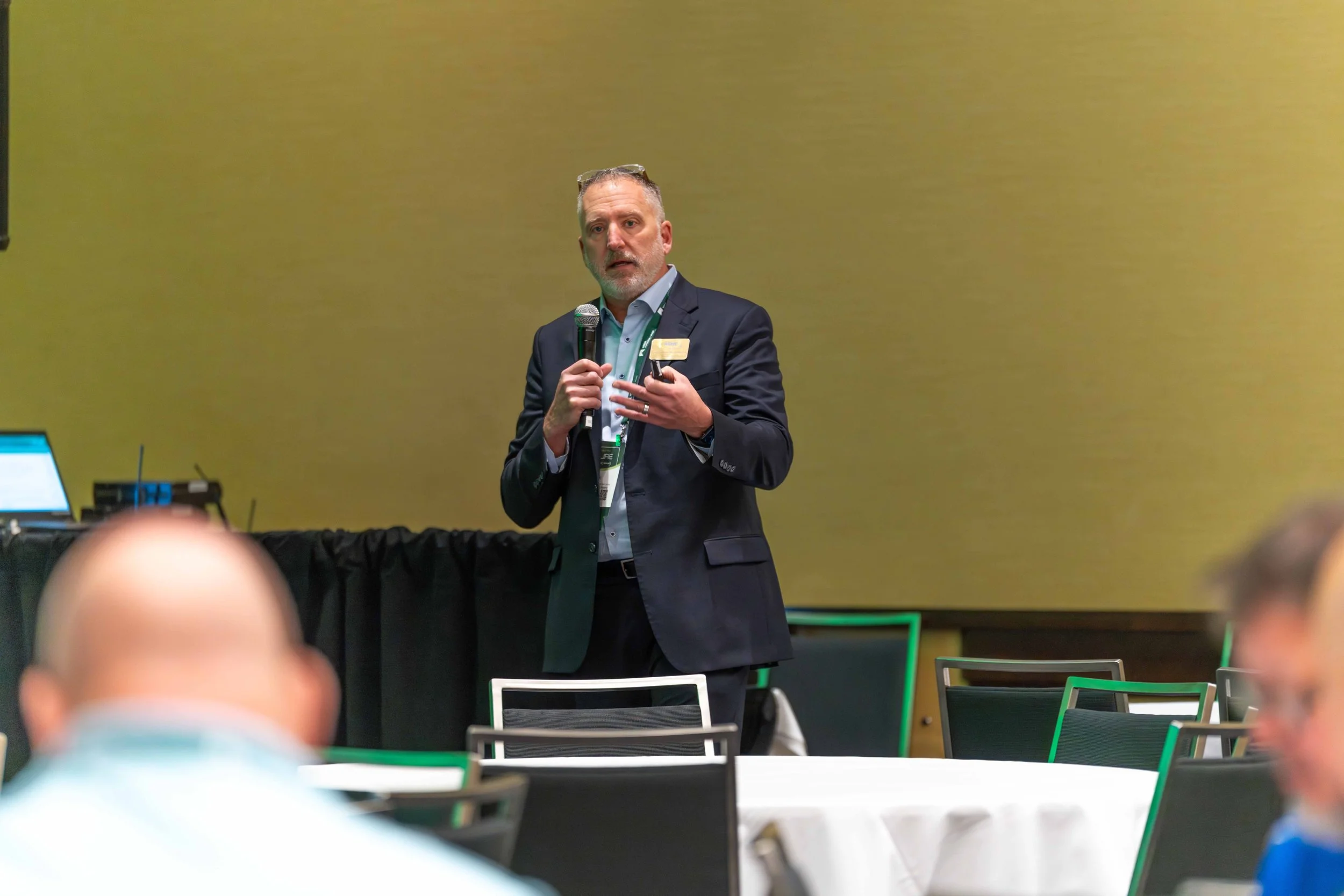A man in a dark suit giving a presentation at a conference, holding a microphone and gesturing with his hand, with room attendees visible in the foreground.