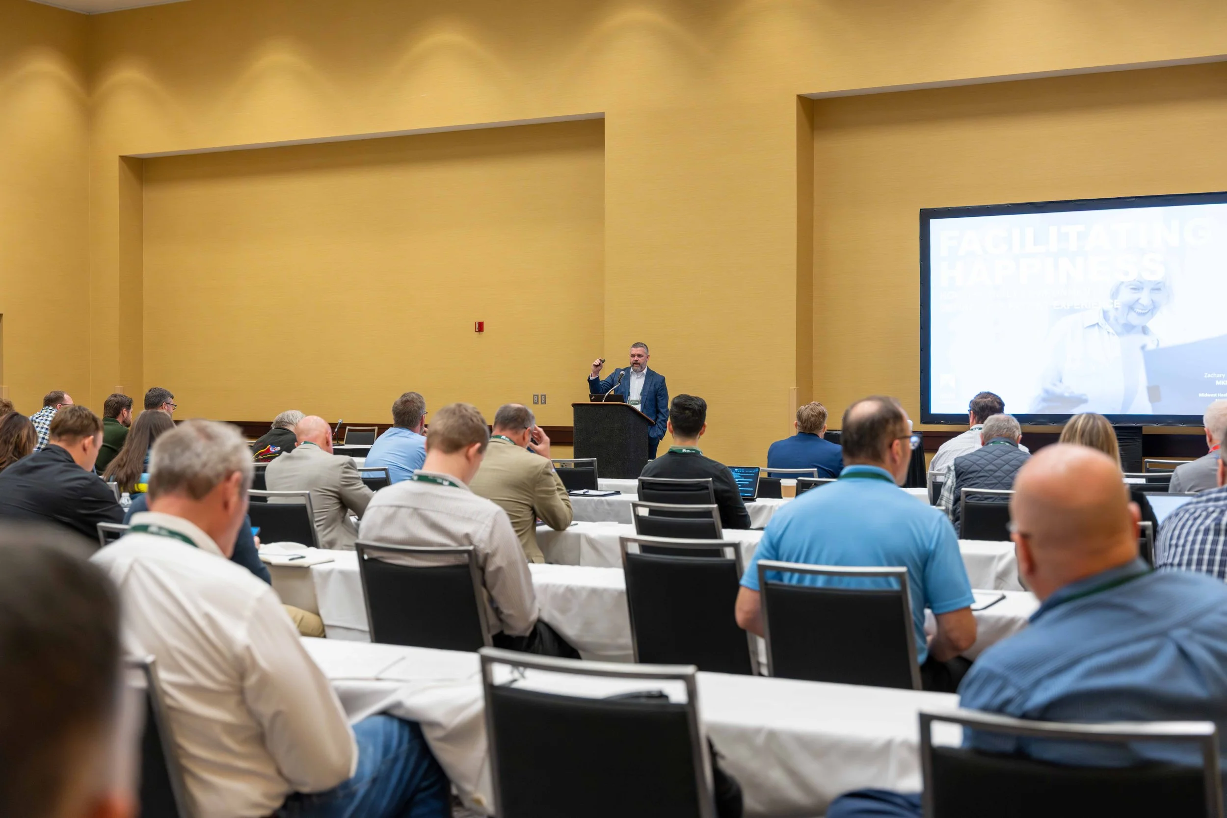 A man in a suit giving a presentation at a conference. The audience is seated at tables with laptops, and a large screen displays a slide with the words "Facilitating Happiness." The conference room has yellow walls and is filled with attendees.