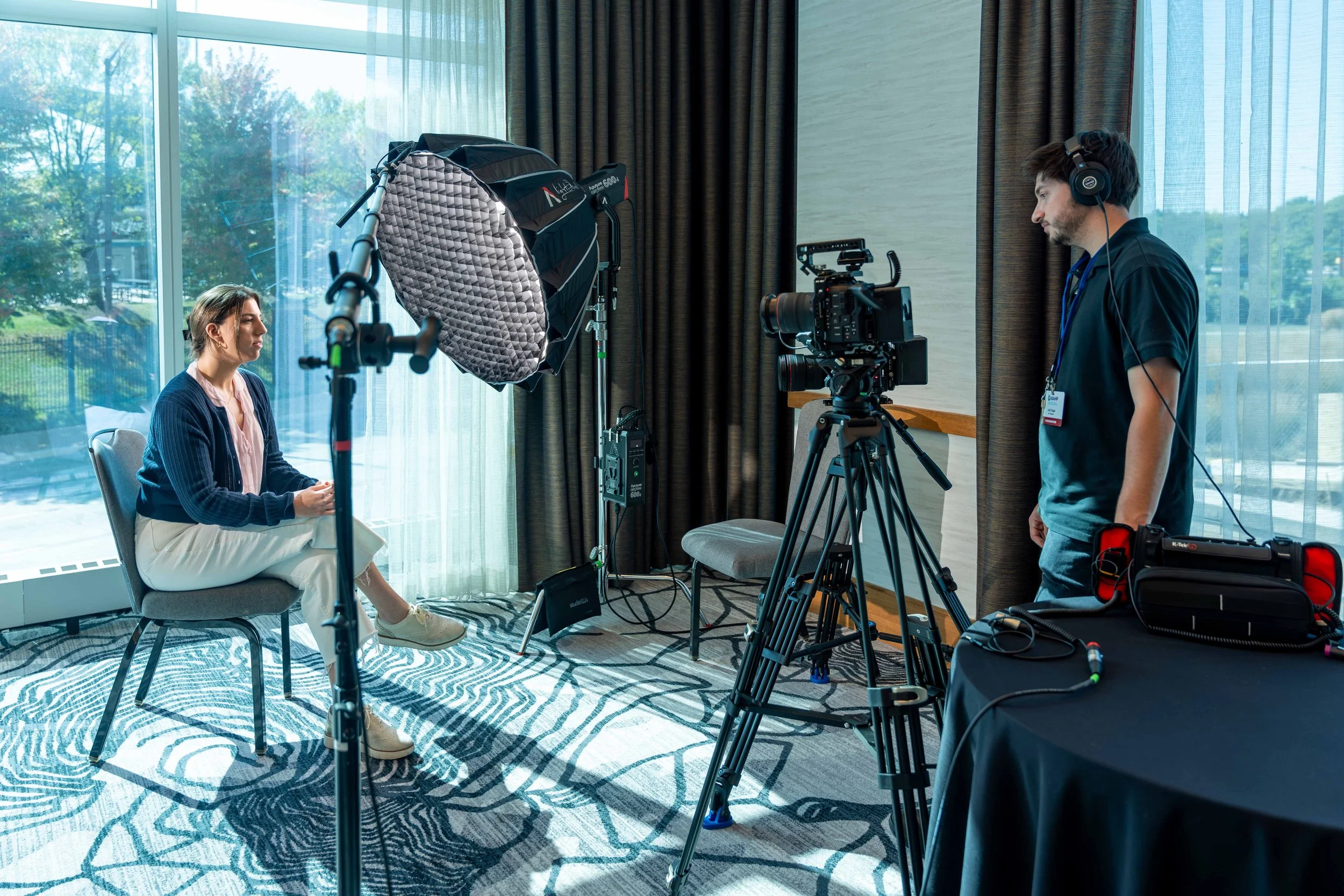 A woman seated facing a camera, during a video recording or interview in a well-lit room with large windows and curtains.