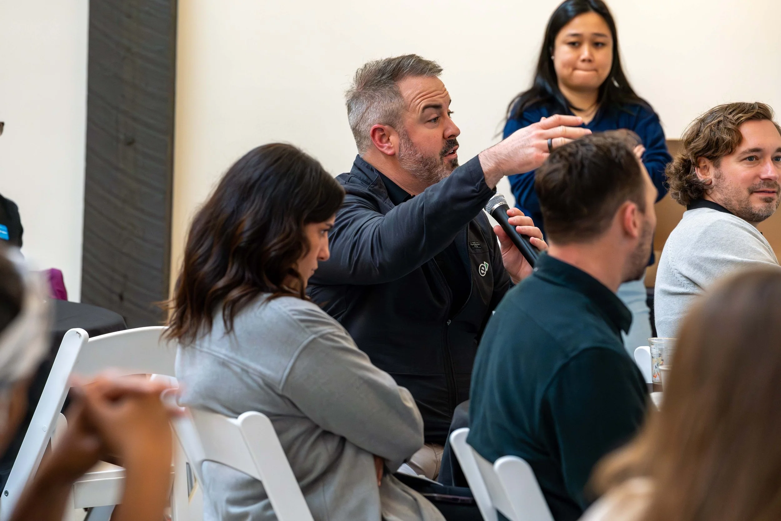A man with gray hair and beard holding a microphone and raising his hand, seated among other people at a discussion or Q&A session.