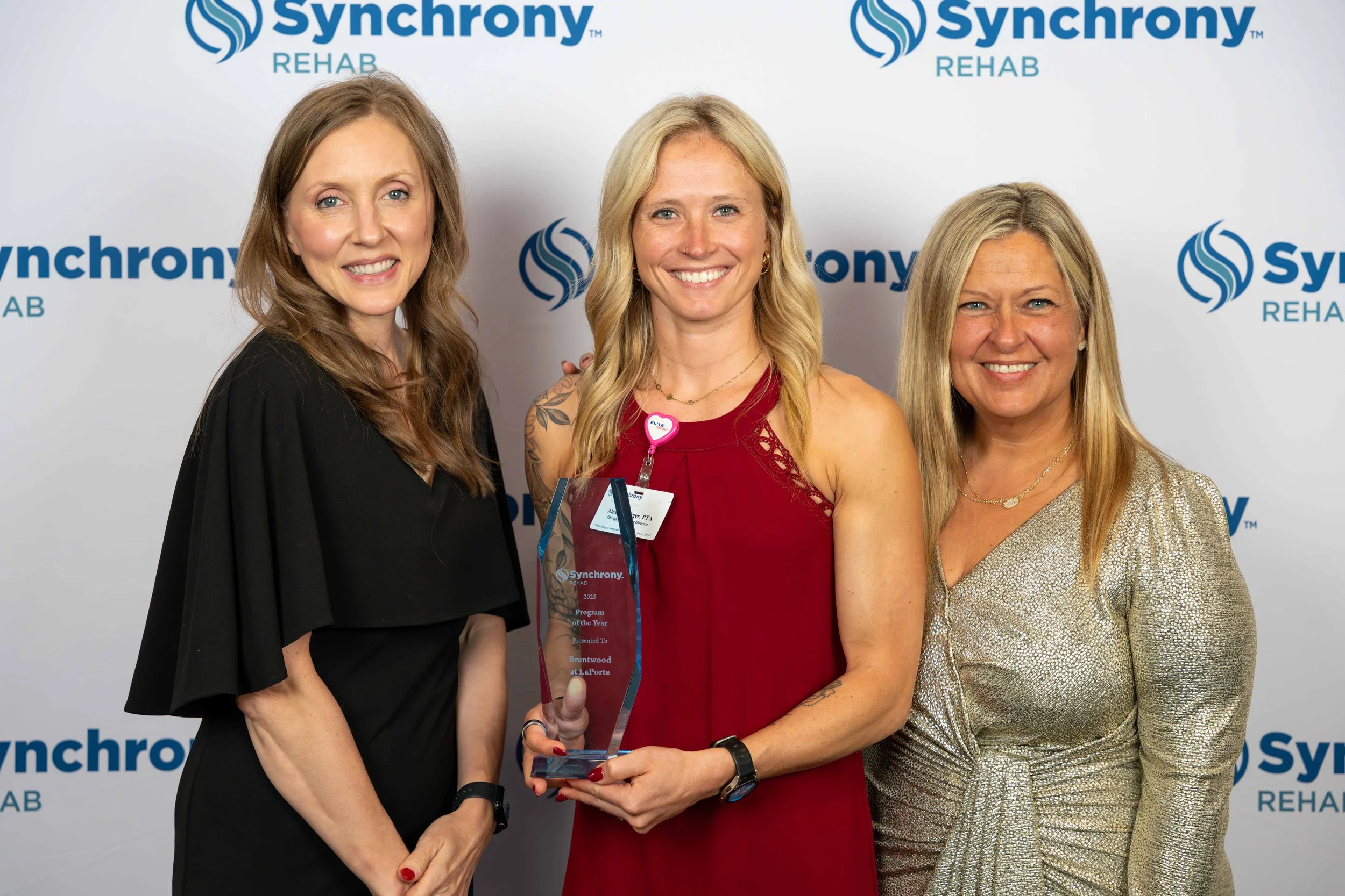Three women at an award ceremony, with a backdrop displaying 'Synchrony Rehab.' The woman in the center, wearing a red dress, holds a glass award and is smiling. The women on either side are also smiling and dressed in elegant attire.