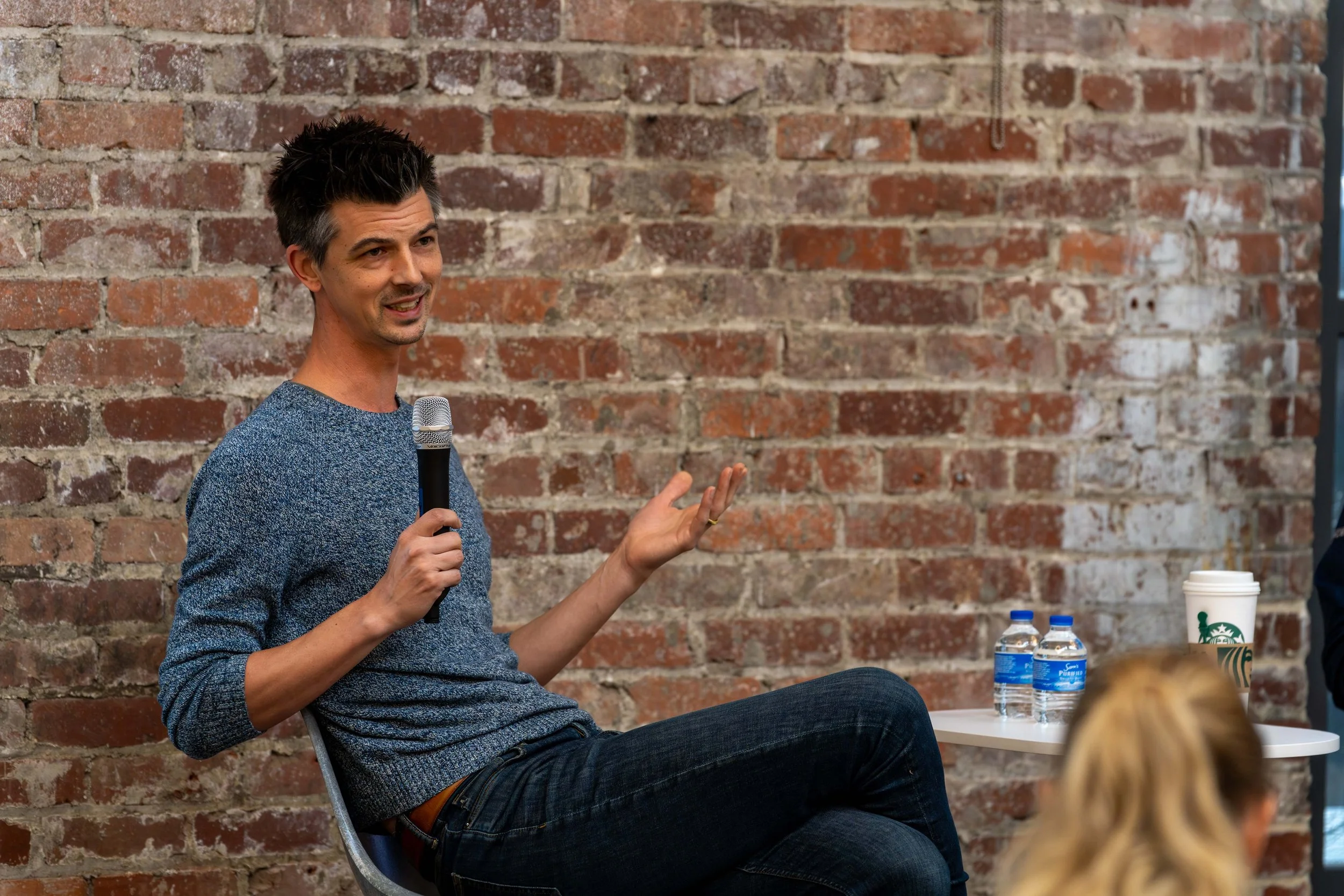 A man sitting on a chair speaking into a microphone during a presentation or discussion, with a brick wall in the background.