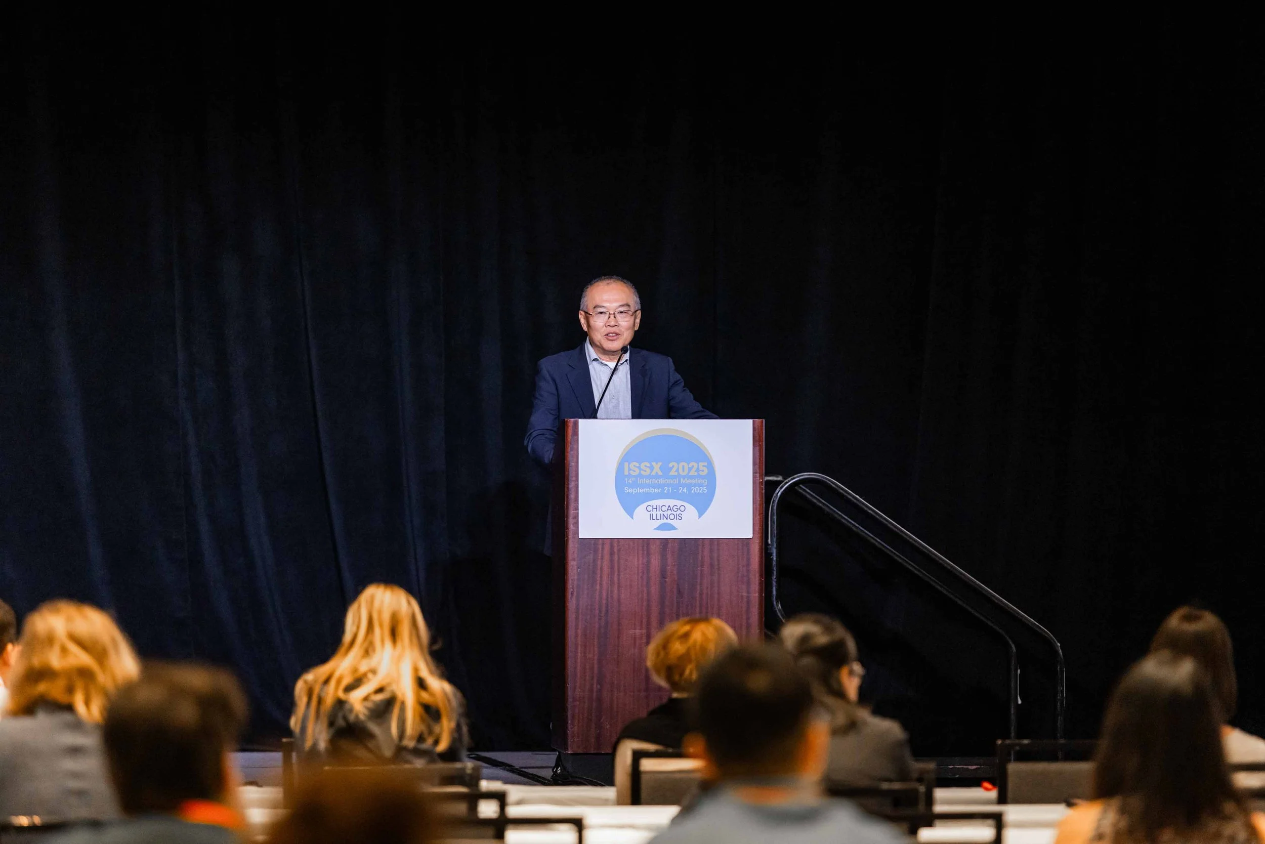 A man speaking at a podium during a conference with a sign that reads ISSX 2025 and Chicago, Illinois, with an audience seated in front of him.