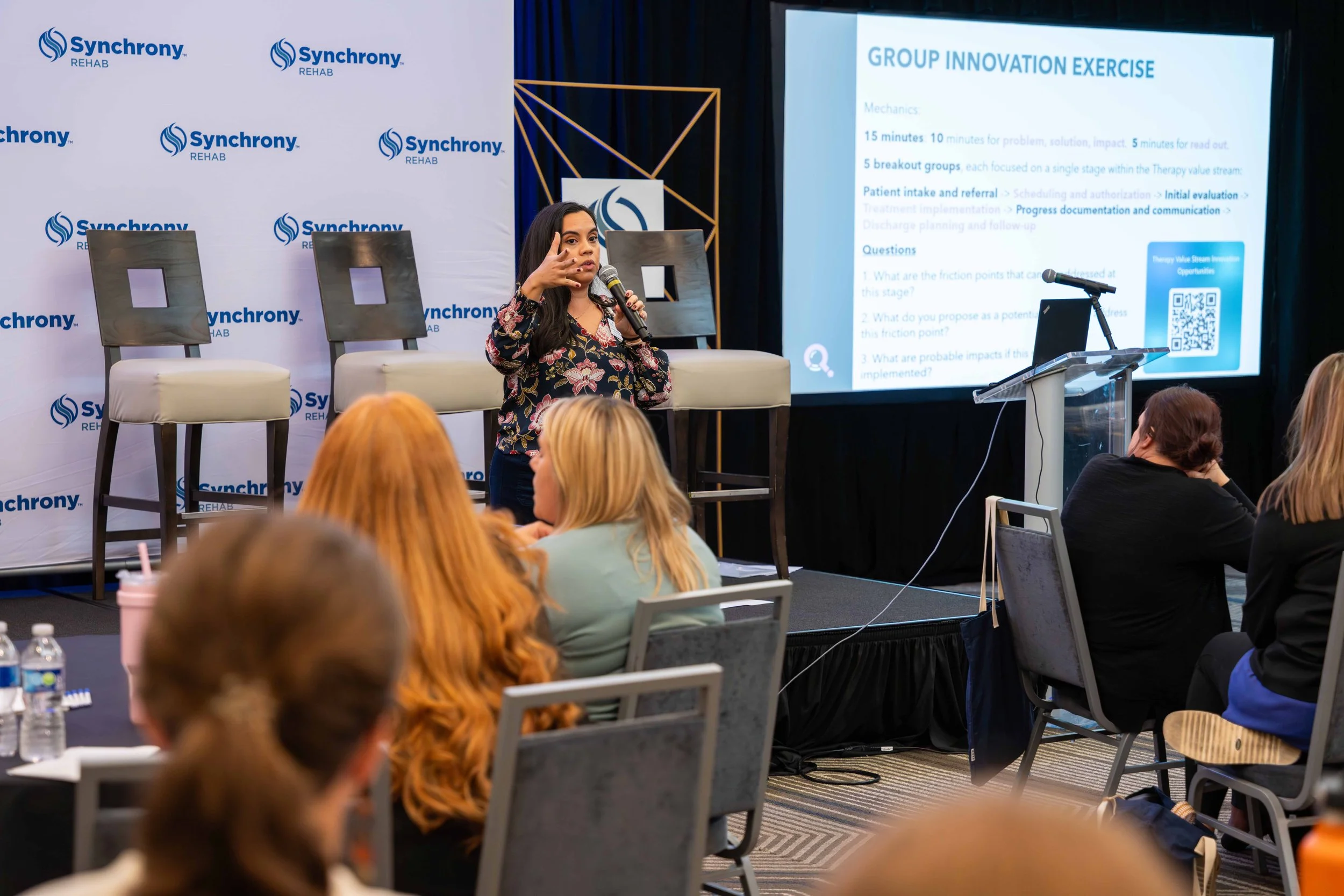 A woman in a floral blouse giving a presentation at a conference with a screen displaying a slide titled 'Group Innovation Exercise' and the logo 'Synchrony Rehab'. Audience members are seated at tables, listening.