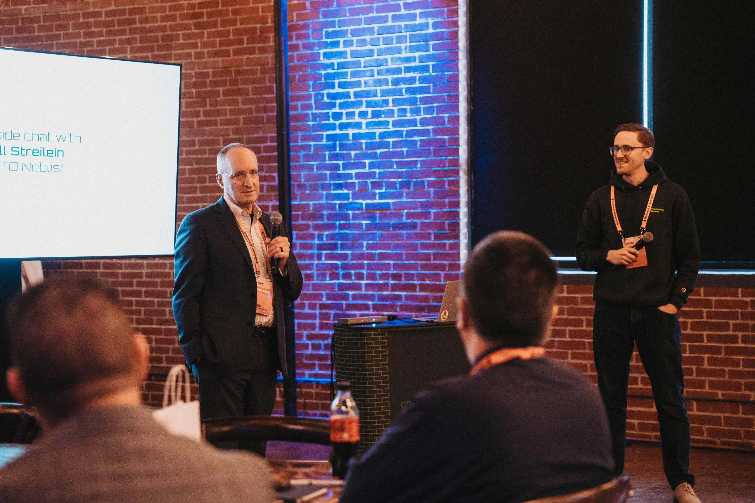 Two men standing on a stage, one speaking into a microphone, the other smiling and holding a microphone, in front of an audience in a room with a brick wall and a large screen display.