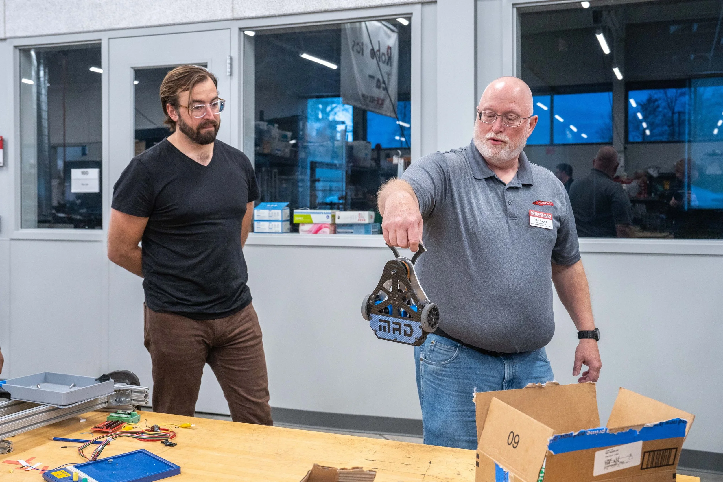 Two men in a workshop or laboratory; one man with glasses, a beard, and dark T-shirt, observing another man who is bald, with glasses, and wearing a gray polo shirt, holding a mechanical device or part.