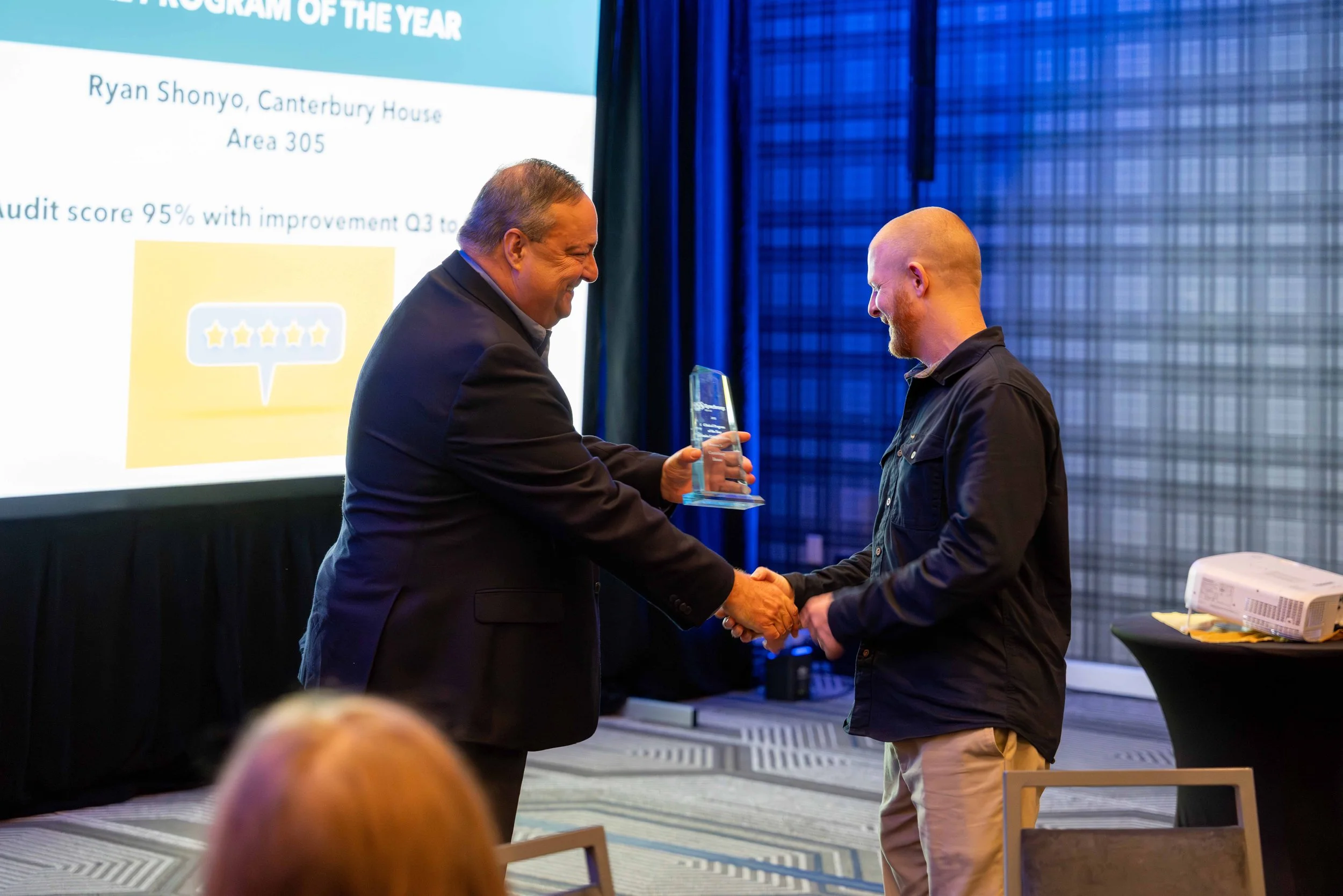 Two men shaking hands during an award ceremony, one presenting a glass award to the other. In the background, a large screen displays a presentation slide with the text 'Program of the Year' and 'Ryan Shonyo, Canterbury House.'