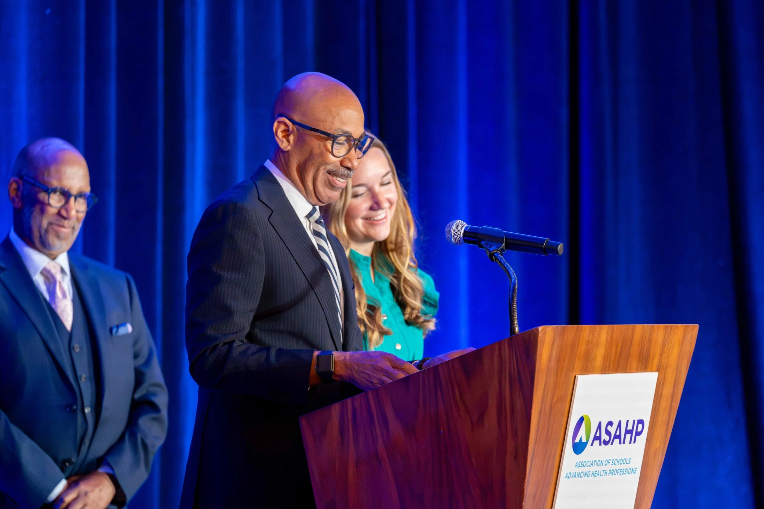 Three people at a podium during a conference with a blue curtain backdrop. The man in the middle is speaking at a podium with ASAPH logo, wearing glasses, a dark suit, and a striped tie. The woman beside him and the man to his left are smiling and dr