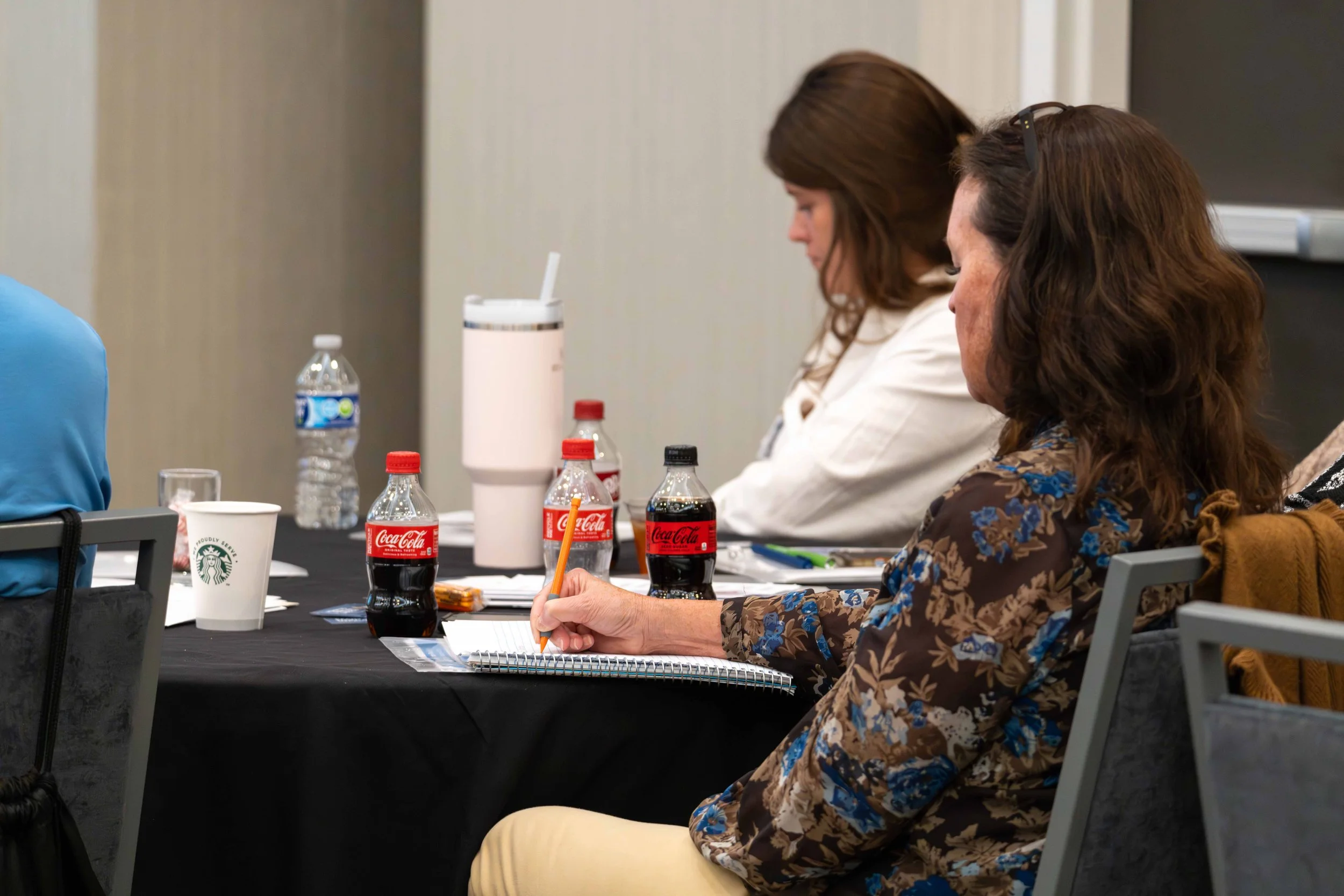 People attending a meeting, take notes, with Coca-Cola bottles, Starbucks cup, and water on the table.