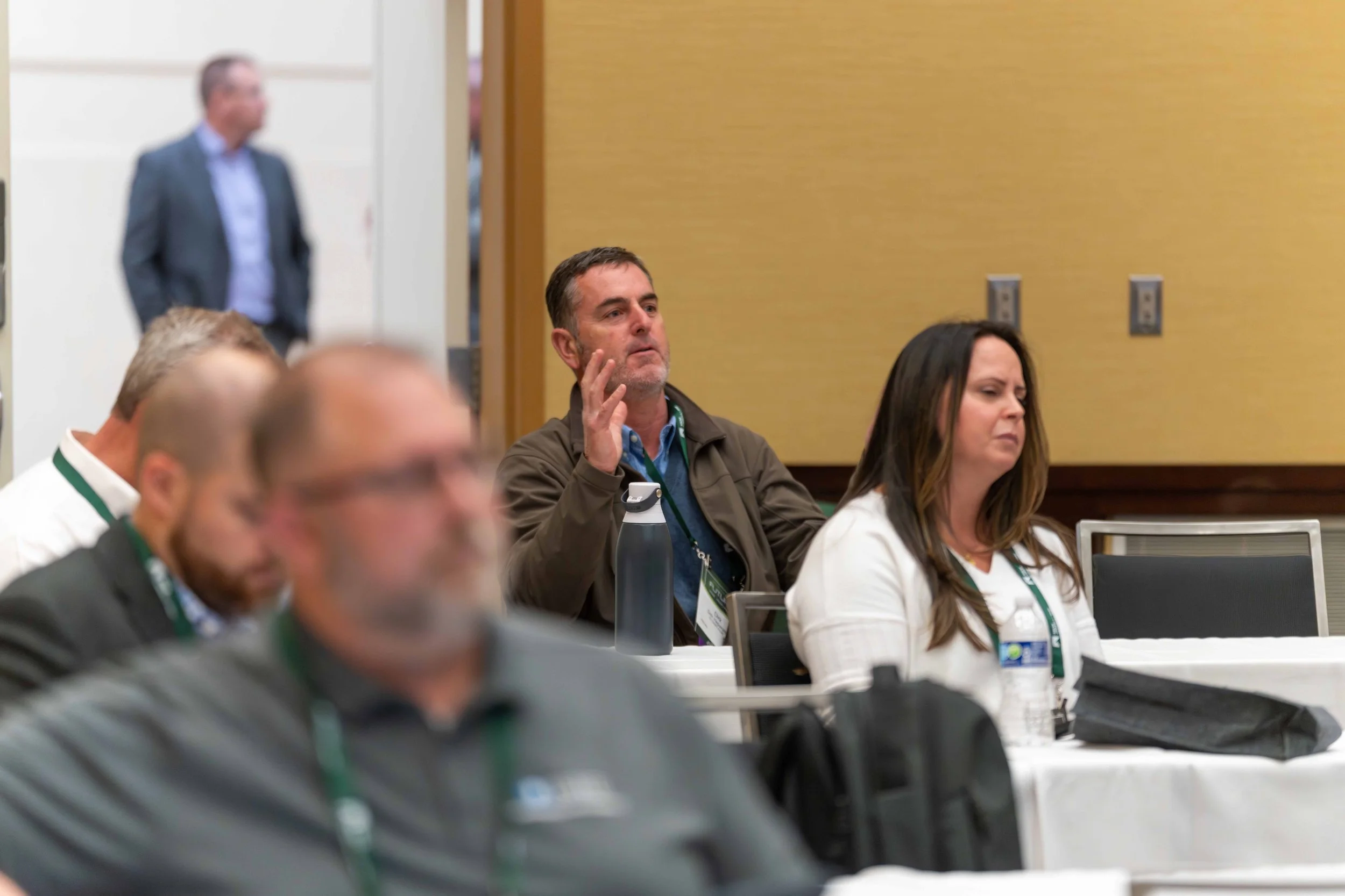 Attendees at a conference, seated at tables, with one man raising his hand and a woman listening, in a room with beige walls.