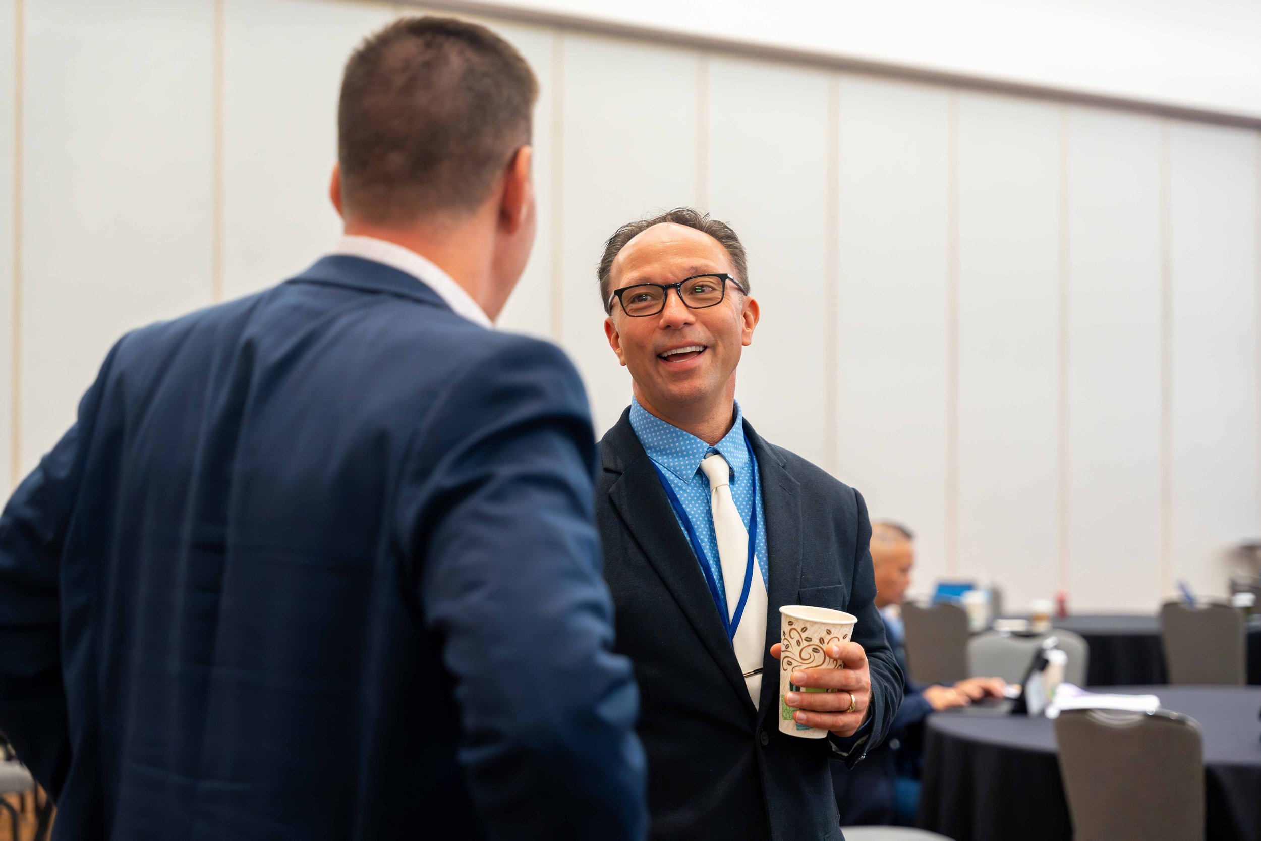 Two men in business suits having a conversation at a professional event, with the man on the right holding a coffee cup and smiling.