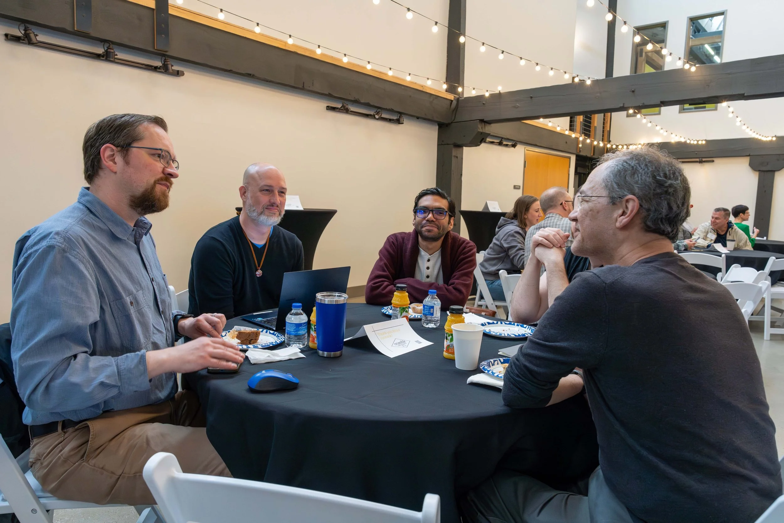 Group of five people seated around a table at an indoor event, with some eating and engaging in conversation, while others are working on laptops, in a well-lit room decorated with string lights.