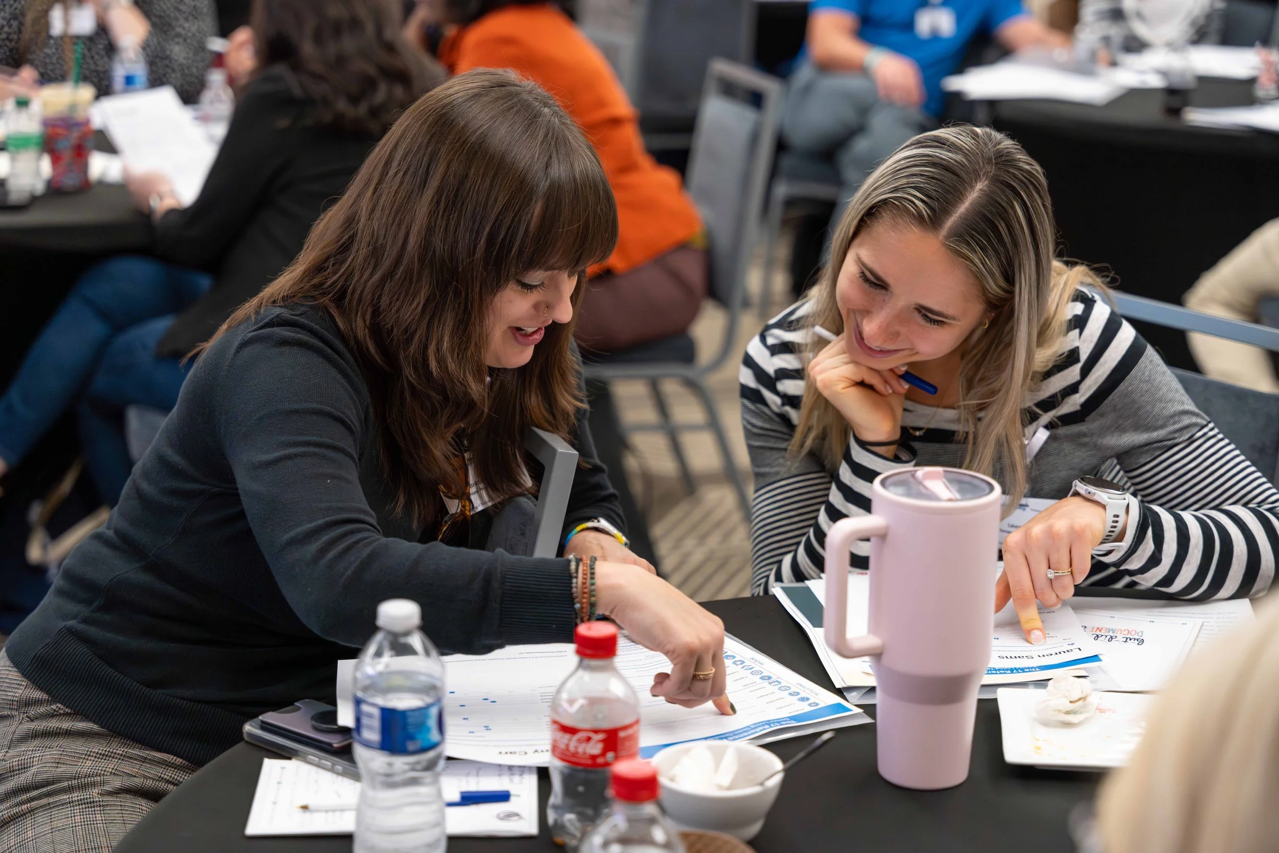 Two women sitting at a table, looking at documents and smiling, in a busy conference or workshop setting with other attendees around.