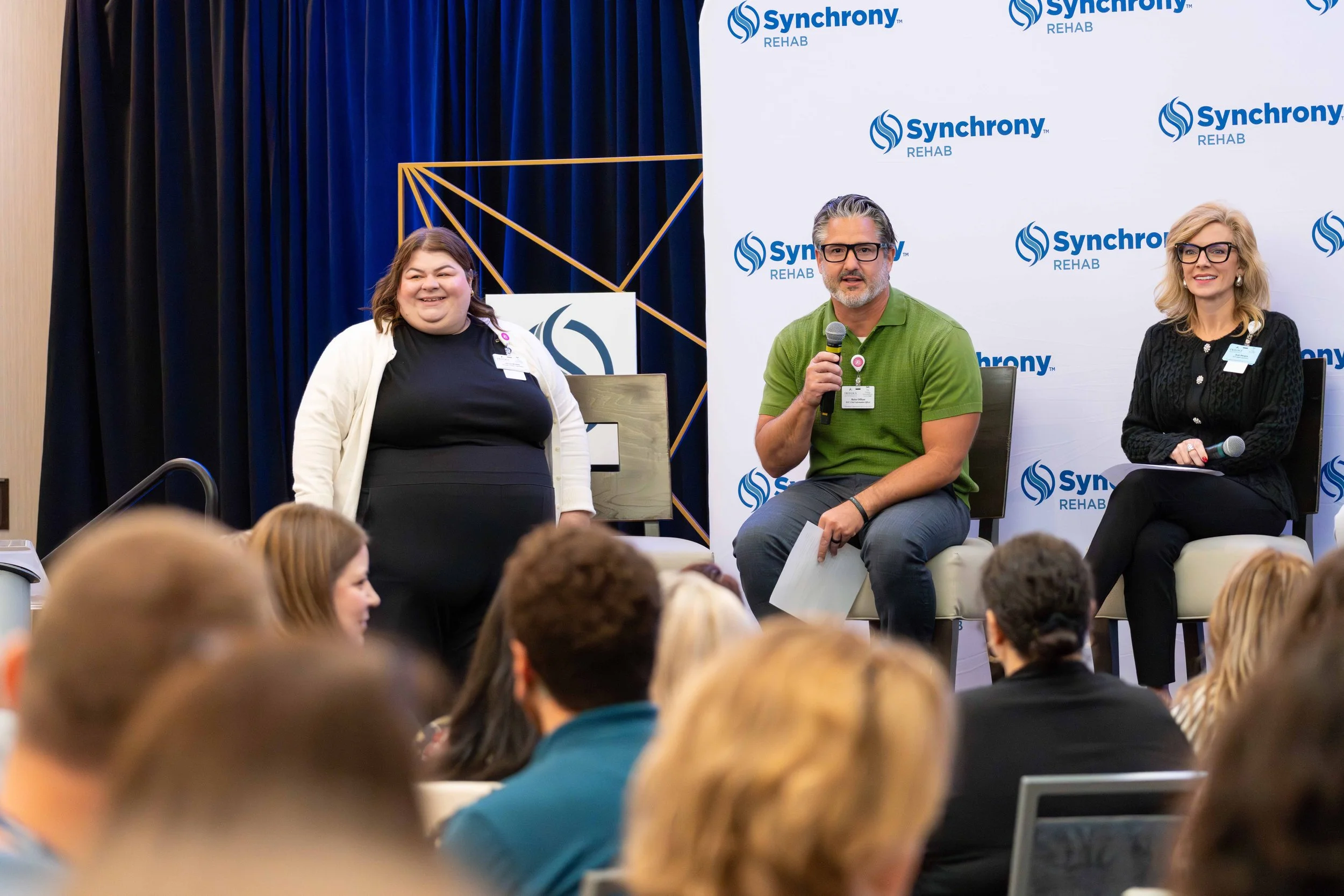A panel discussion at a conference with three speakers on stage, two sitting and one standing. The backdrop displays the 'Synchrony Rehab' logo. The audience is attentively listening.
