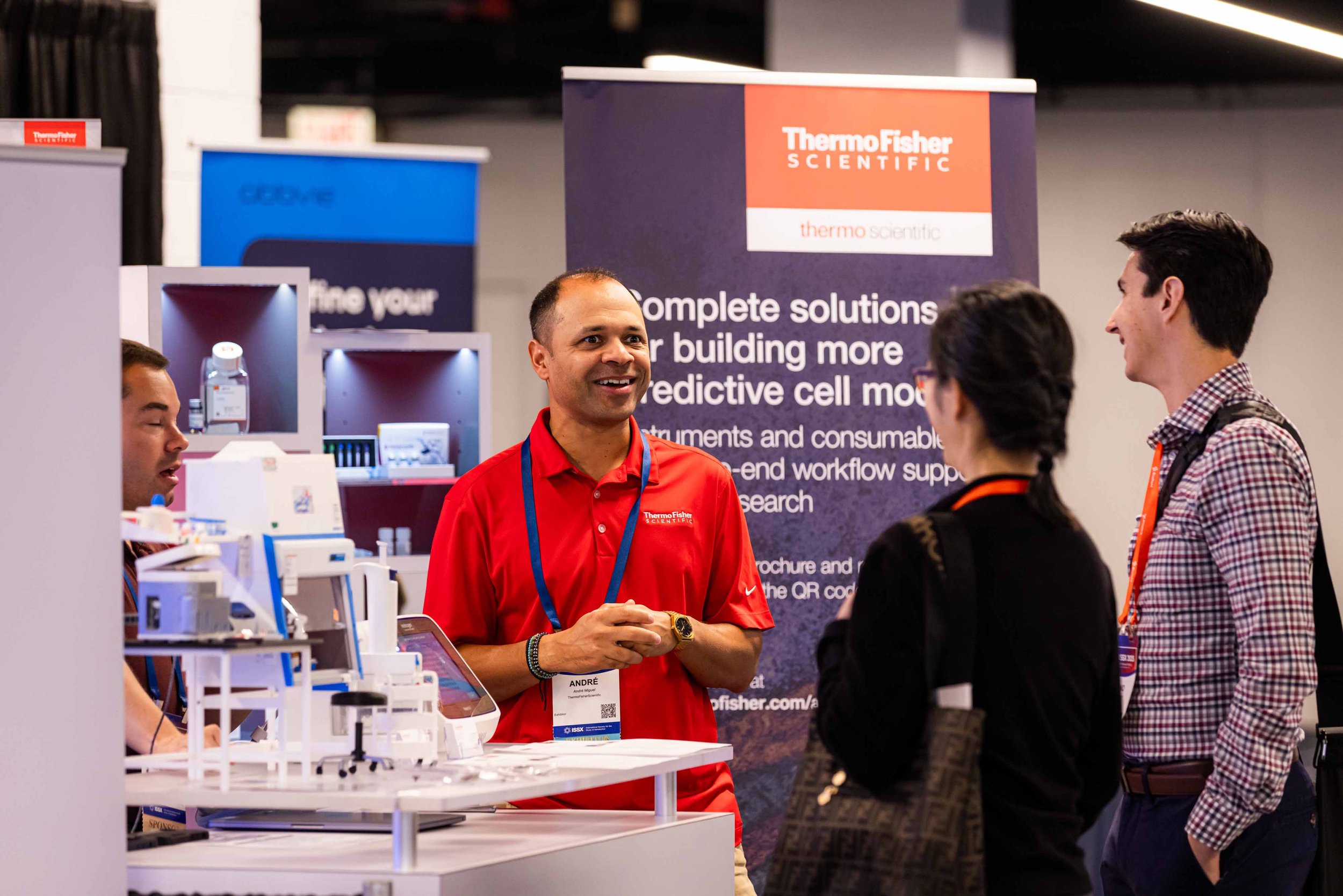A man in a red shirt at a trade show booth talking to two visitors, a woman and a man, with scientific equipment and branding for Thermo Fisher Scientific in the background.