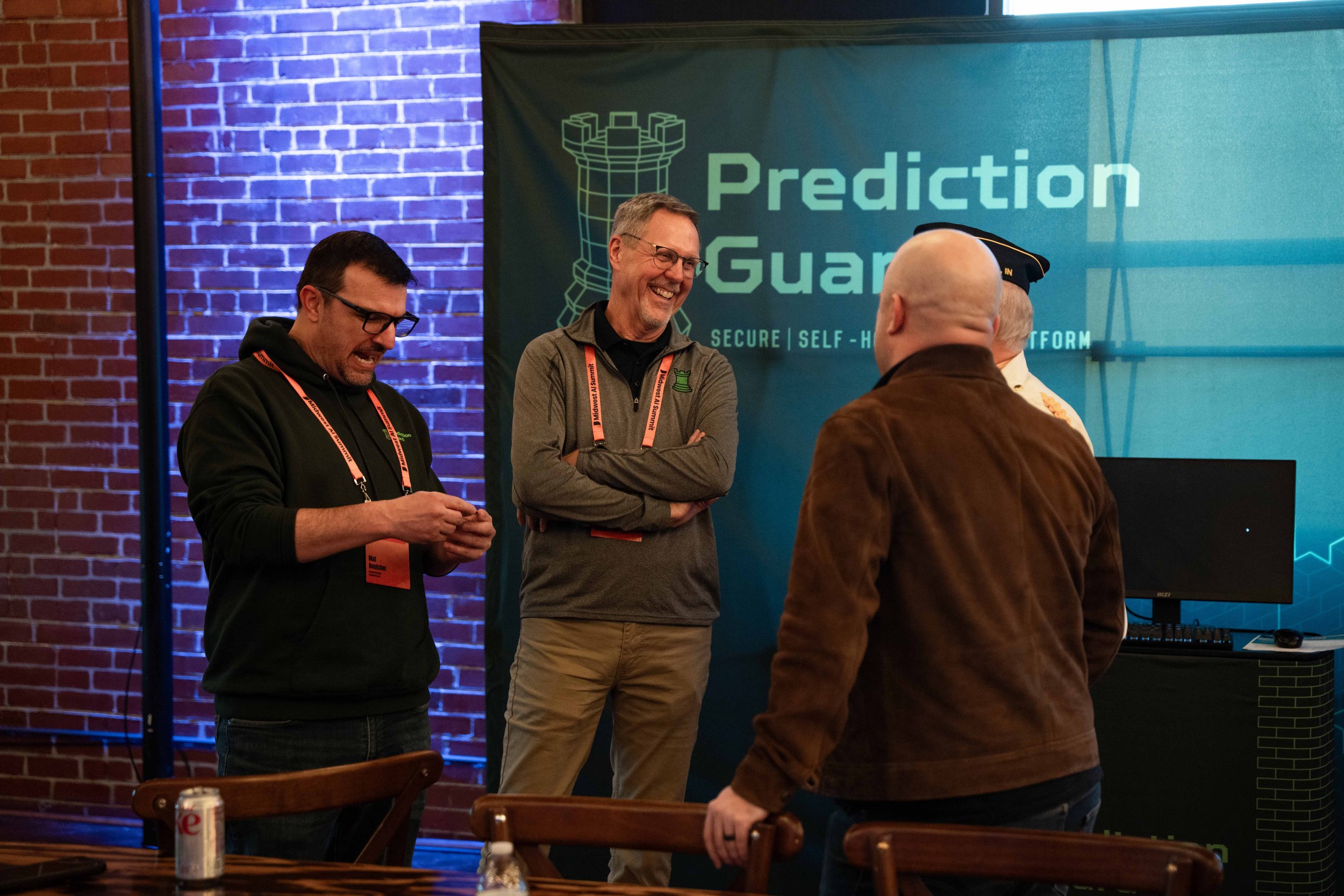 Four men are engaged in a conversation at a tech conference, standing near a blue and green banner that reads 'Prediction Guard' with a chess rook icon. The background includes a brick wall, and there is a table with a soda can and water bottle in th