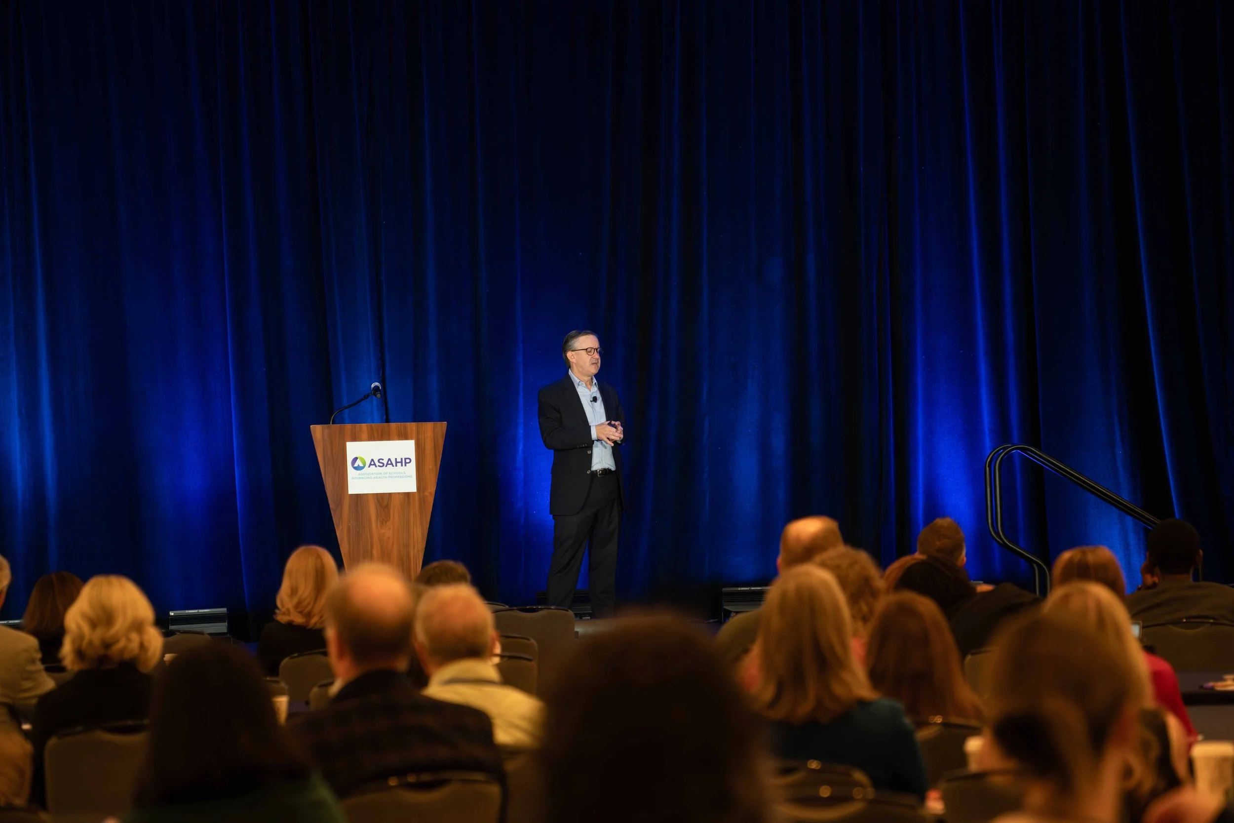 A man in a suit giving a presentation on stage at a conference, with an audience seated in front of him and a blue curtain backdrop.