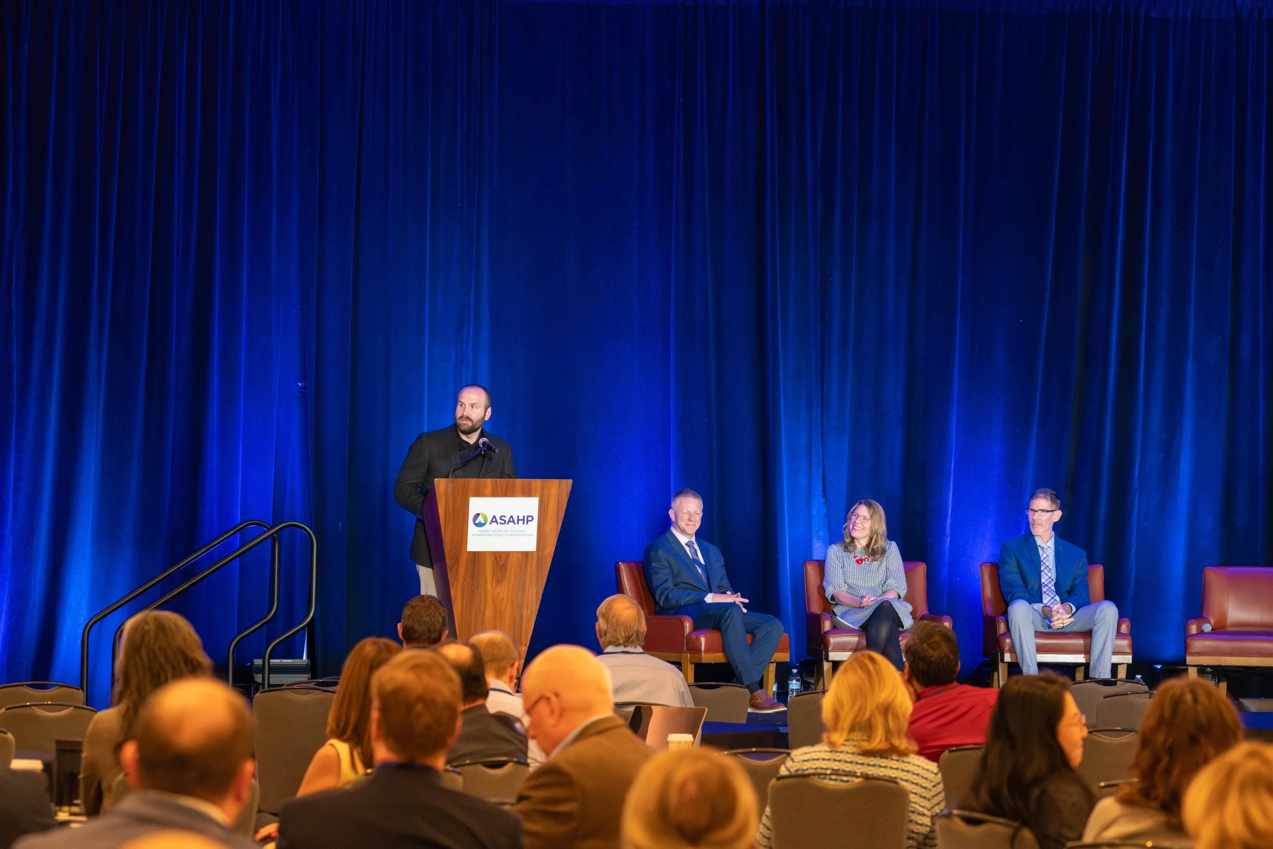 A panel of three people seated on stage during a conference, with a man in a black suit standing at a podium speaking. The background features blue curtains and the podium has a sign with the logo of ASHP. Audience members are seated in front.