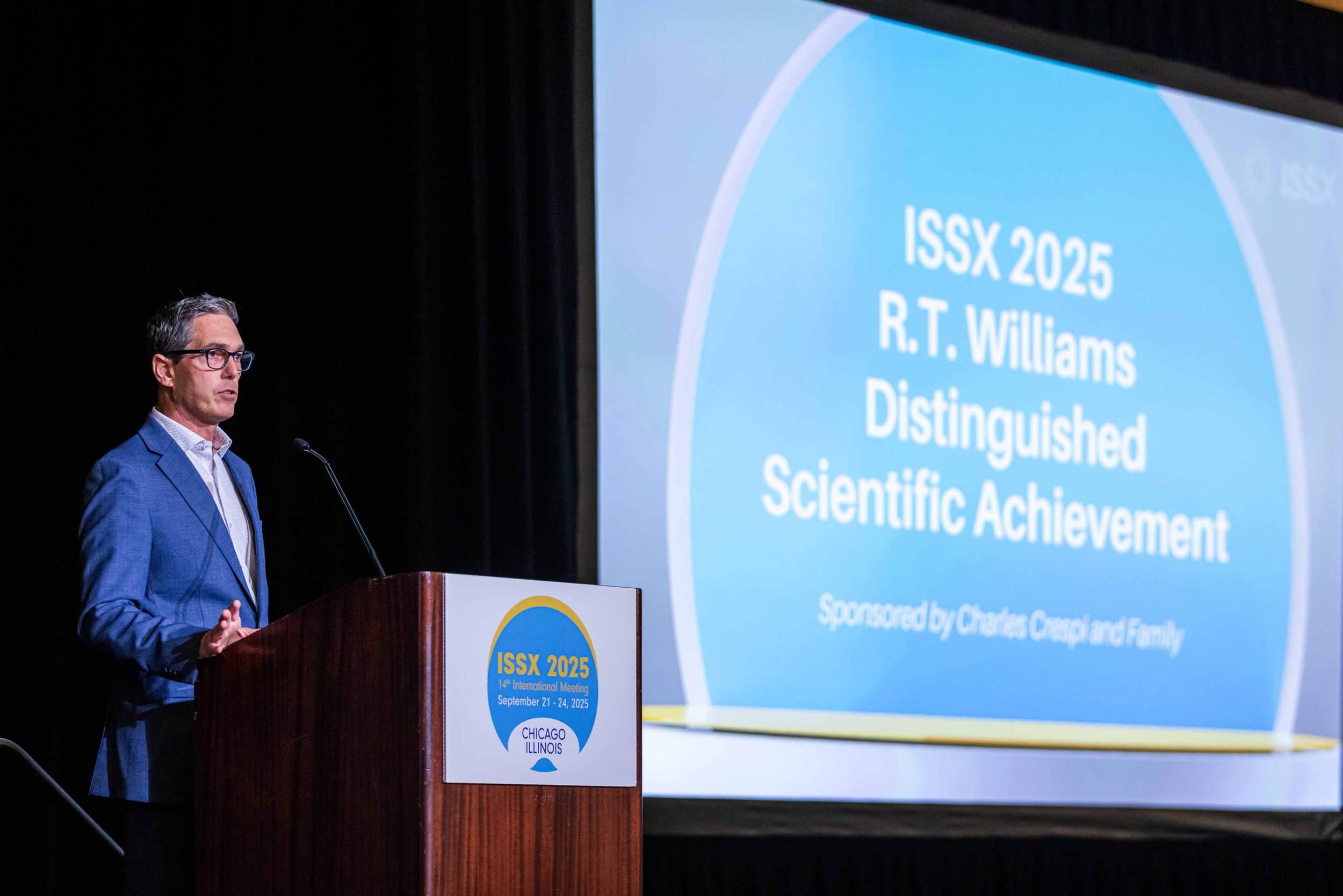 A man in a blue suit and glasses standing at a podium giving a presentation. The podium has a sign for ISSX 2025, the 14th International Meeting, scheduled for September 21-24, 2025, in Chicago, Illinois. A large screen behind him displays the event 