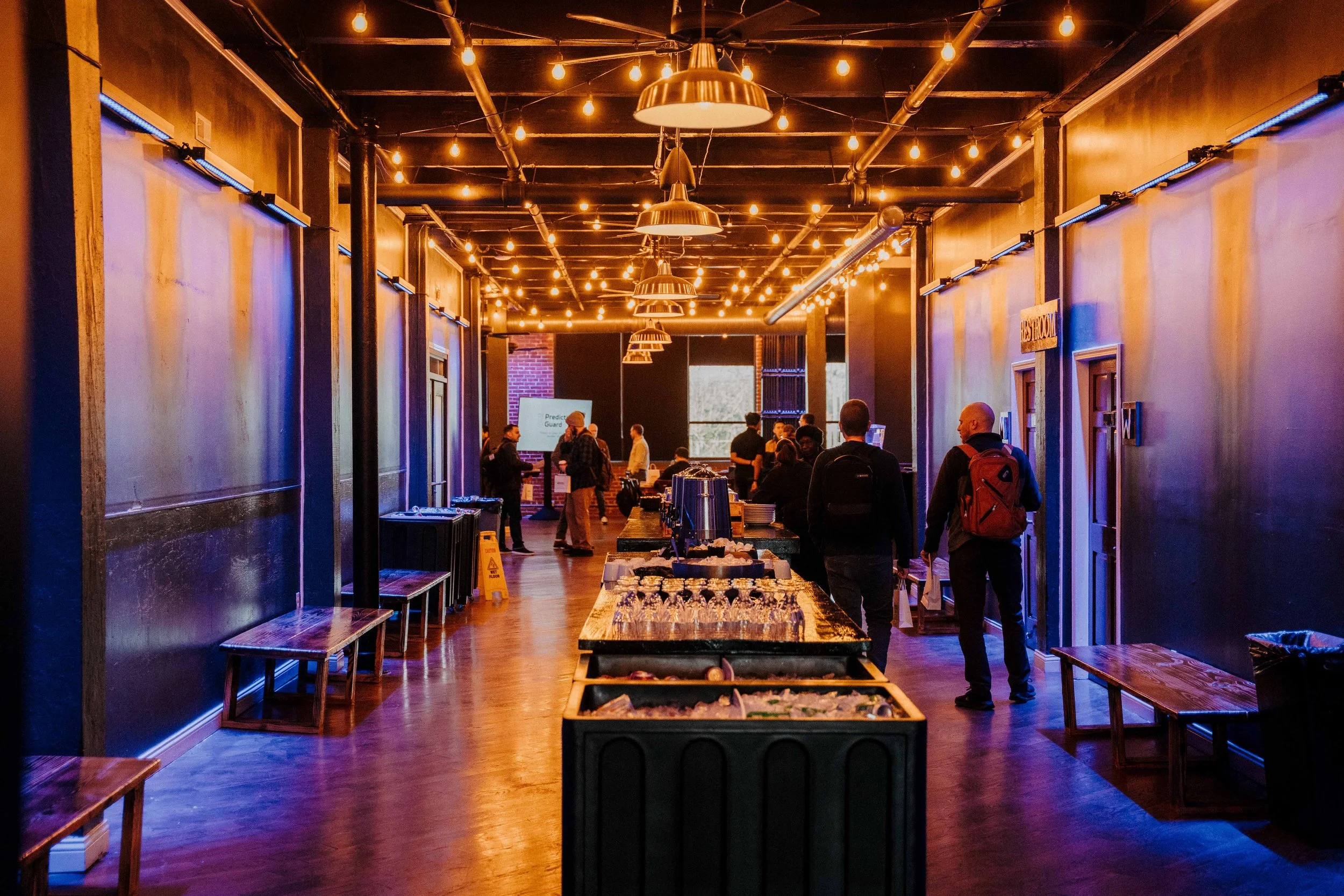 People in a modern event space with string lights, wooden benches, and a long table on a polished wooden floor.