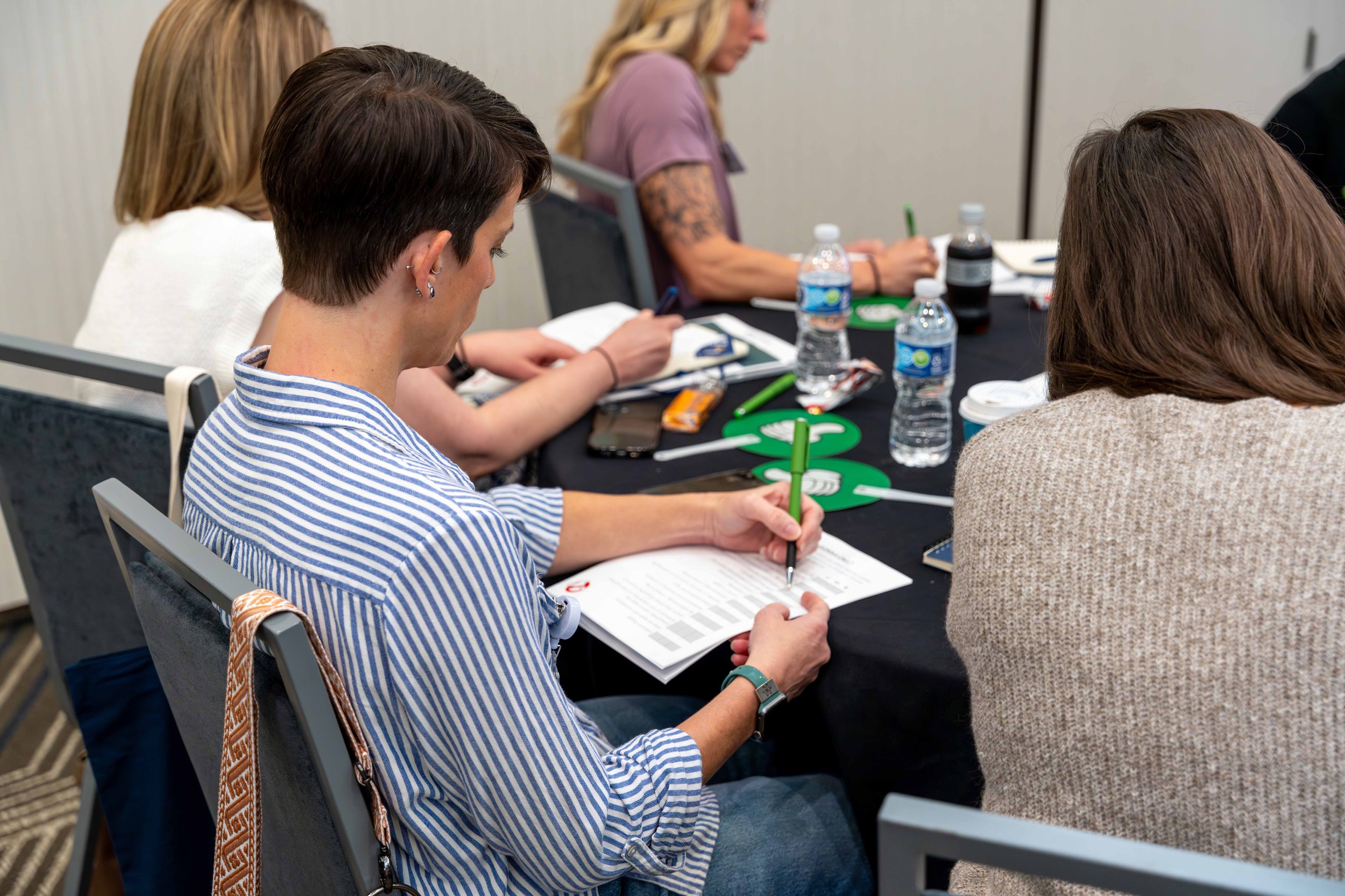 People sitting at a conference table, taking notes with green pens, surrounded by water bottles and notebooks.