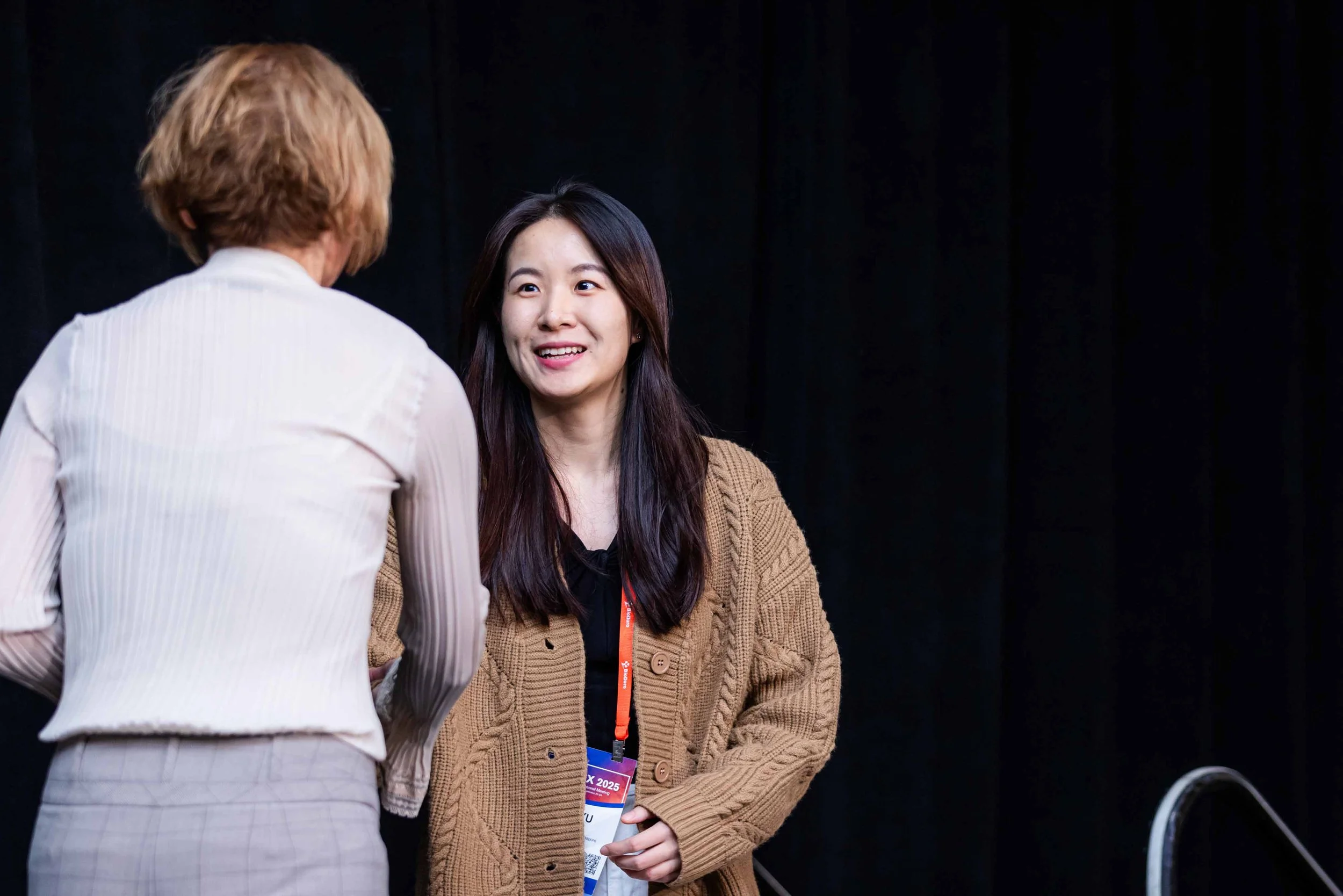 Two women talking, one facing away with short blond hair and a white ribbed top, the other facing forward with long dark hair and a brown sweater, against a black background.