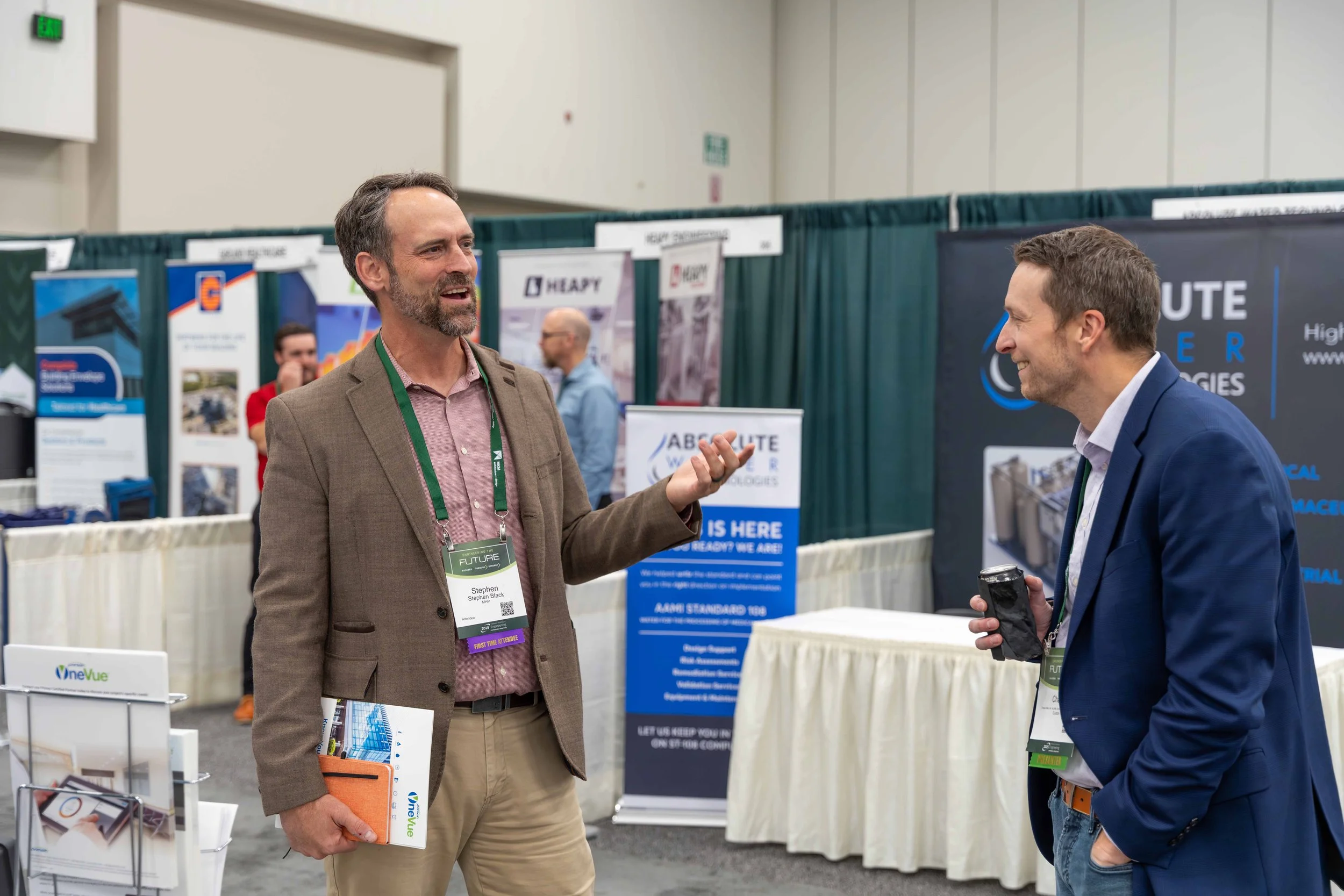 Two men engaged in conversation at a trade show or conference, with booths and banners behind them.