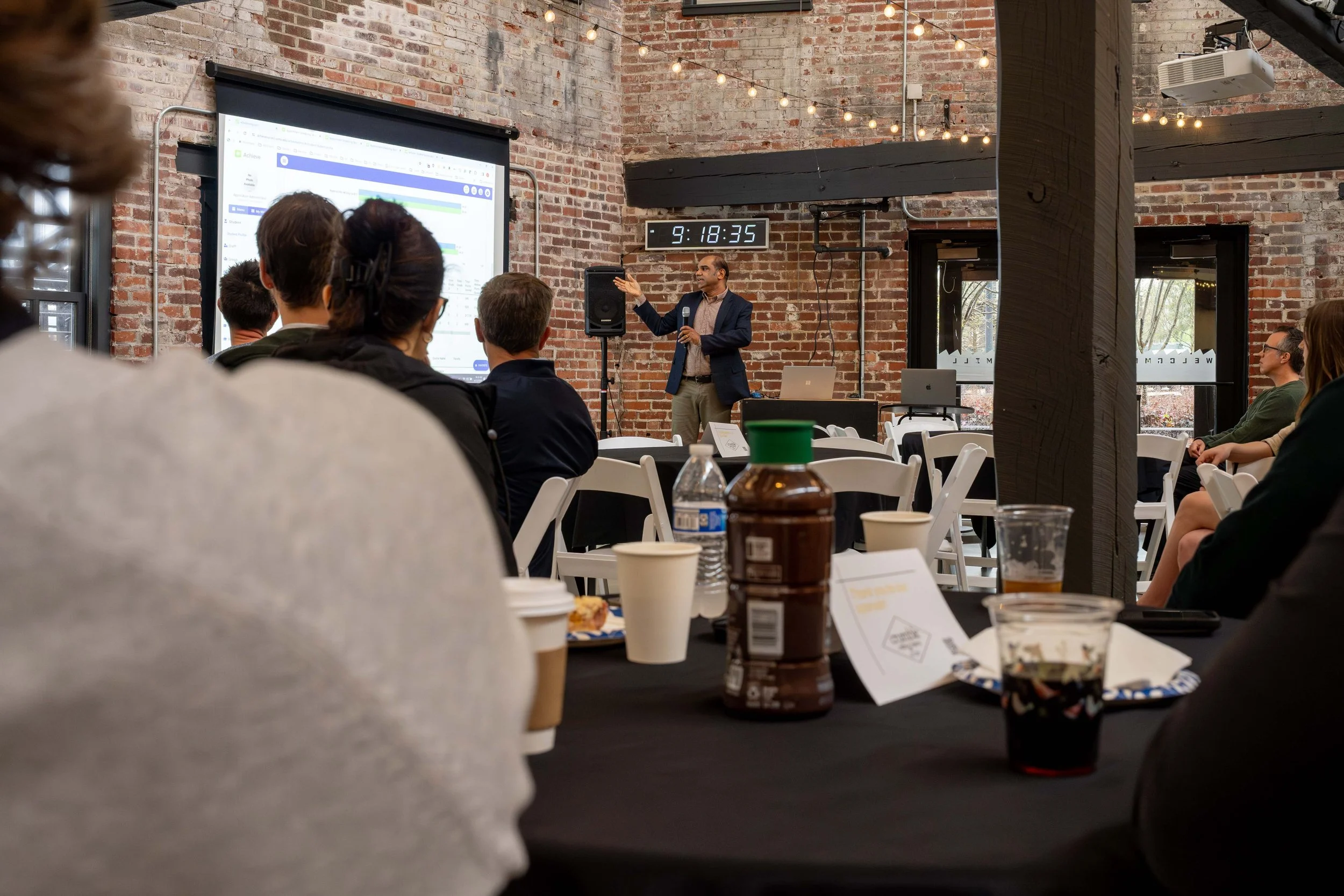 A business presentation in a brick-walled cafe with people seated at tables, a speaker in a suit standing at the front gesturing towards a screen, and a clock showing 9:18:35.