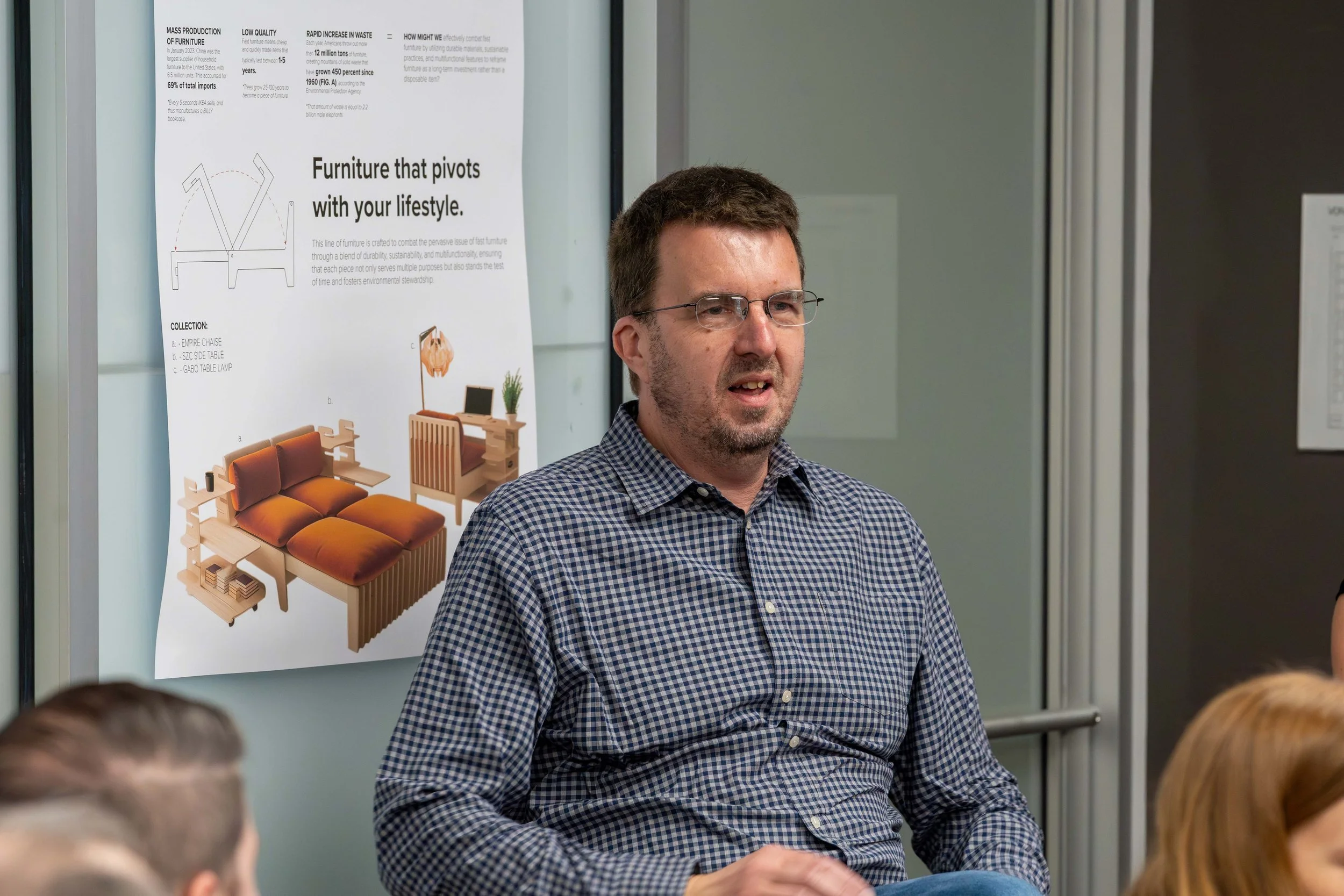 A man with glasses and a short beard, wearing a checkered shirt, appears to be speaking during a presentation. Behind him is a poster about furniture with images of a sofa, chair, and table in a well-lit room.