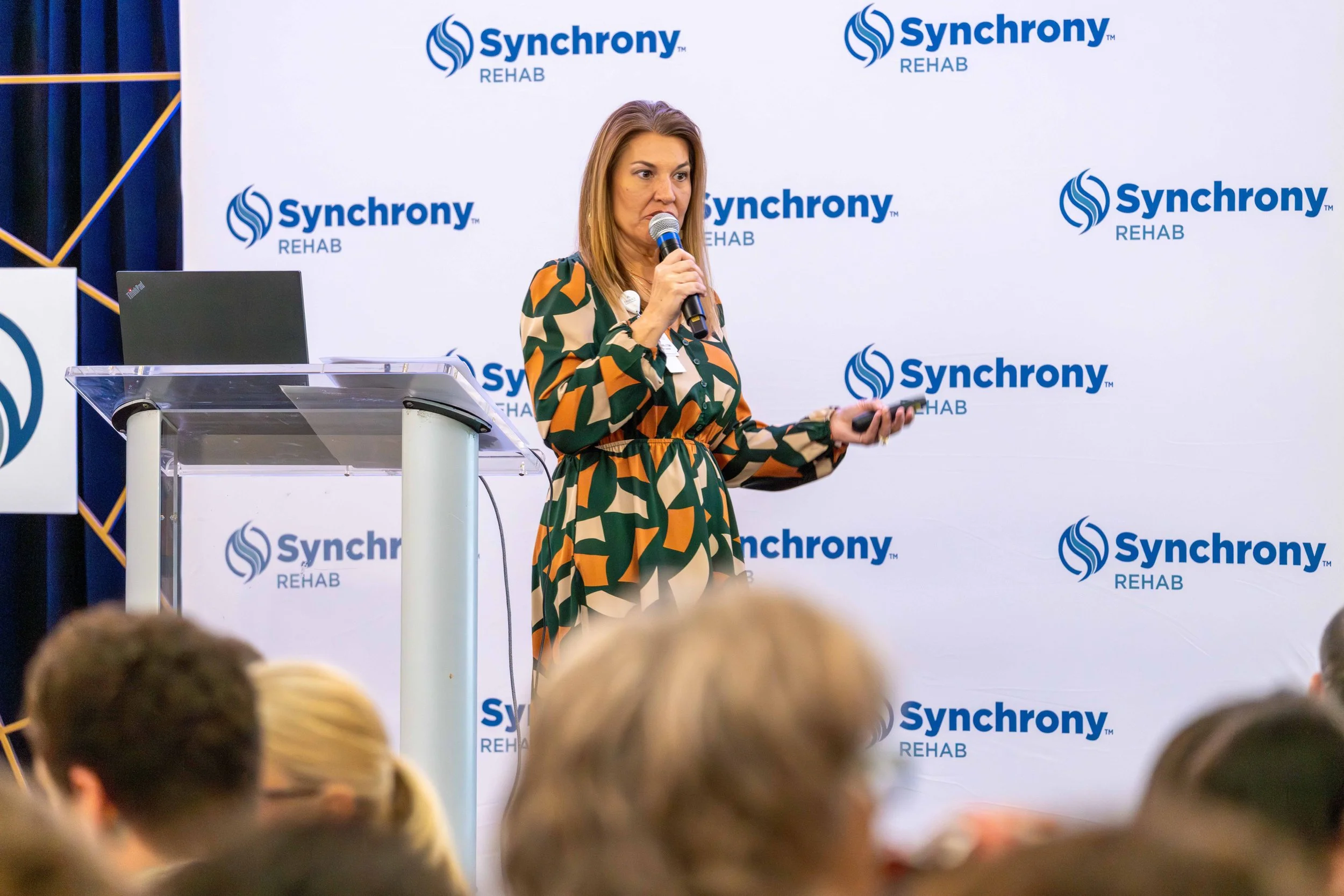 A woman with shoulder-length hair in a colorful patterned dress giving a presentation with a microphone and remote control on a stage with a backdrop that reads "Synchrony REHAB" and an audience in the foreground.