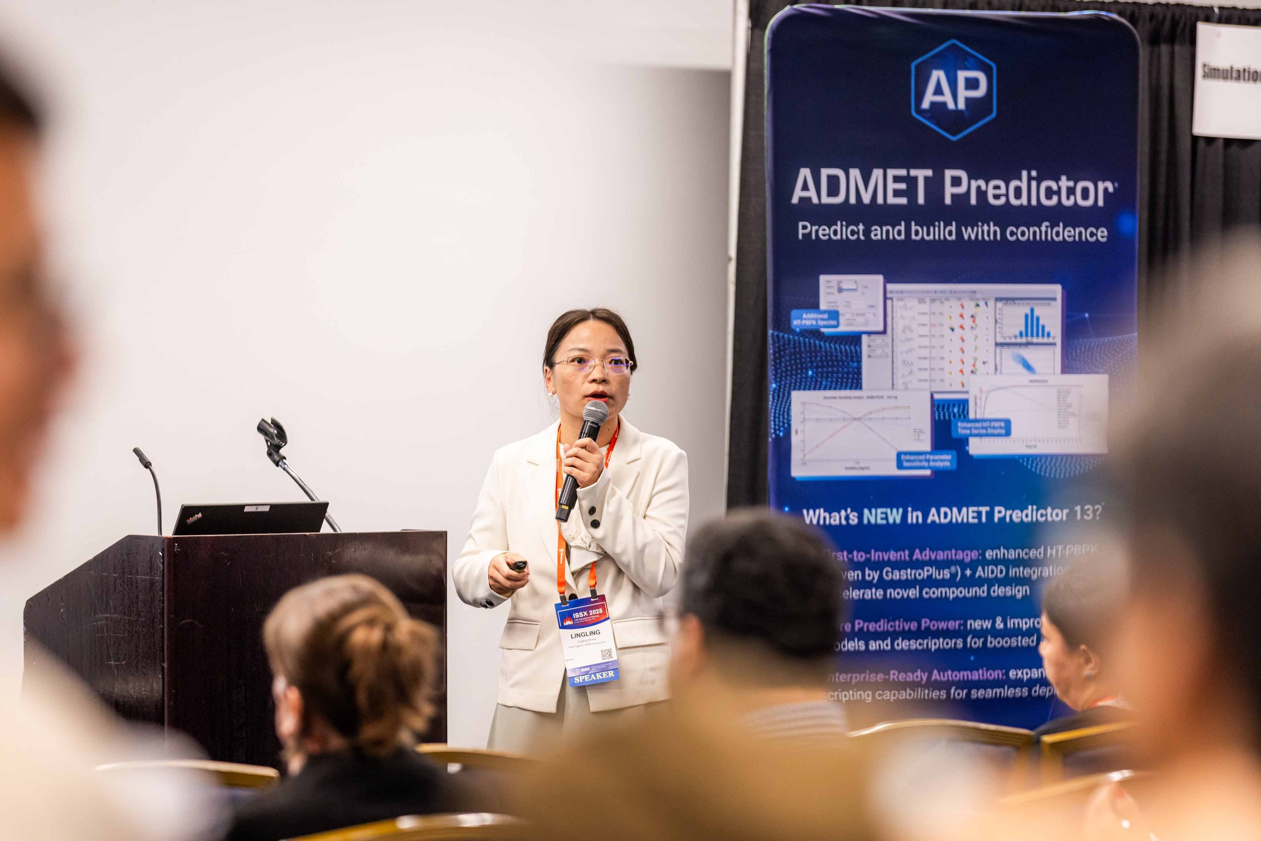 A woman speaking at a conference, standing next to a large banner promoting ADMET Predictor software, with audience members in the foreground.