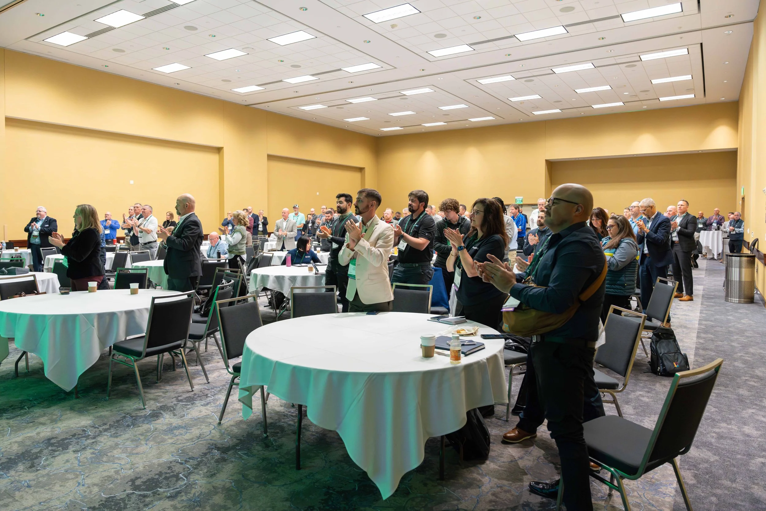 Conference attendees standing and clapping in a large, well-lit room with round tables and beige walls.