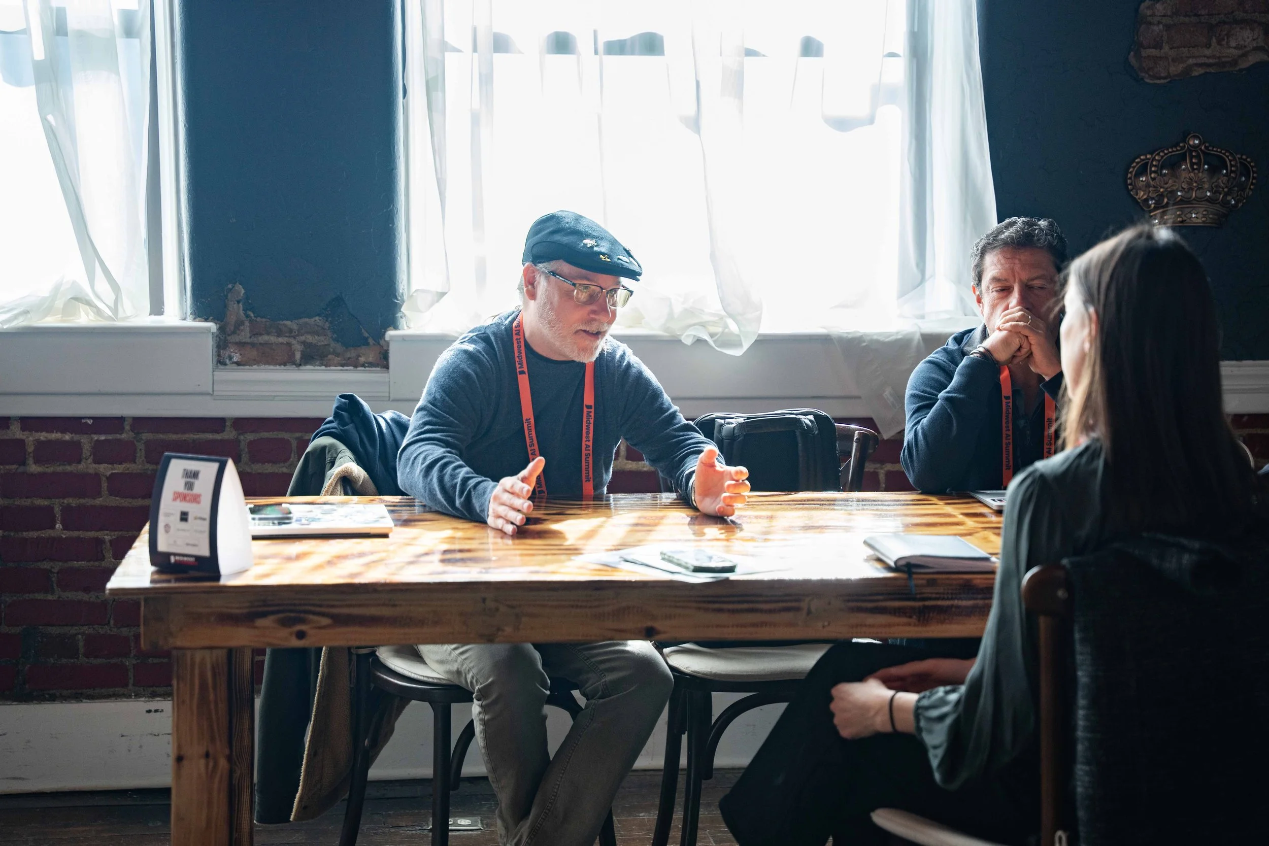 Three people sitting at a wooden table in a room with brick and blue walls and large windows with white curtains, engaged in conversation or discussion.