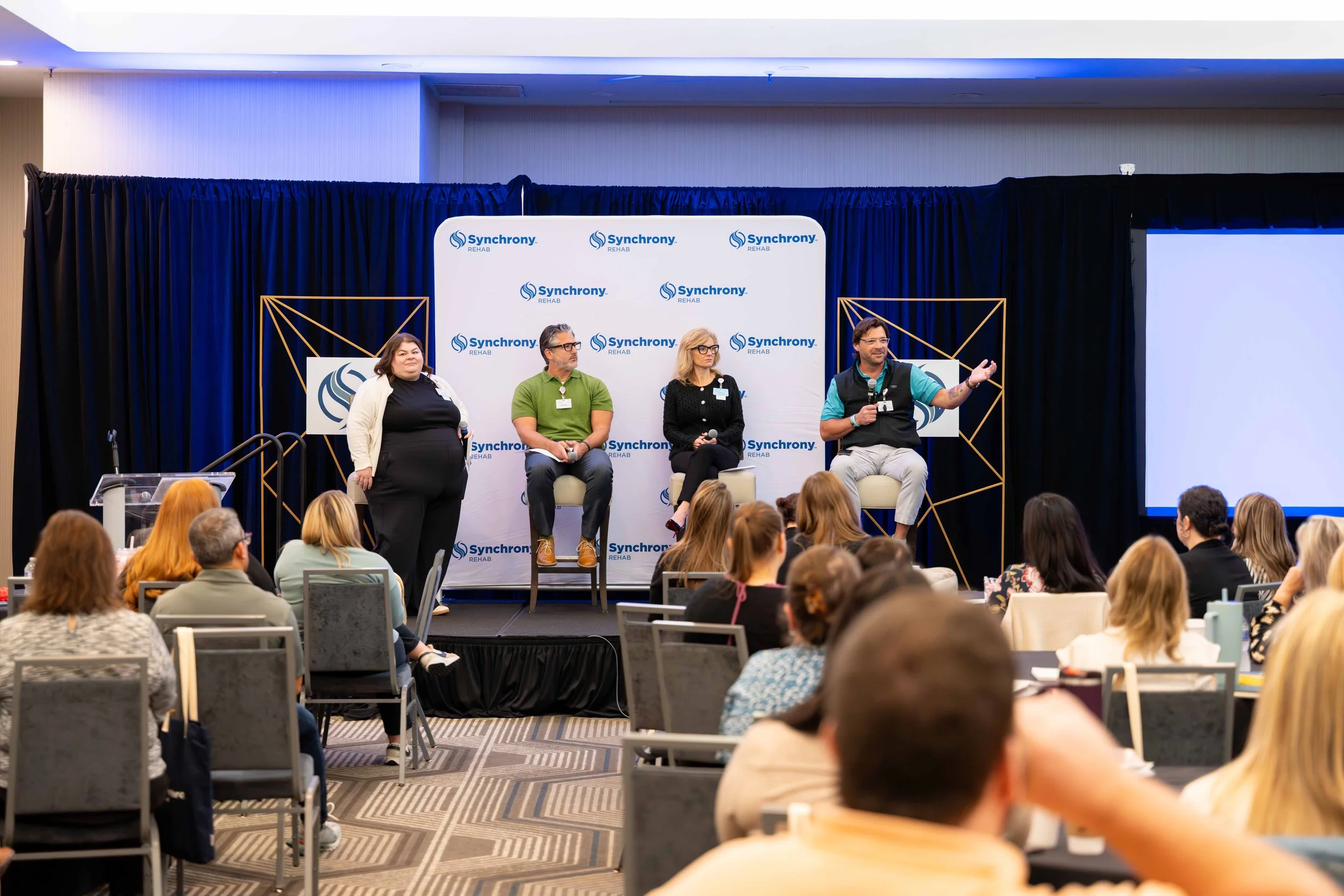 Panel discussion at a conference with four speakers on stage, one speaking into a microphone, and an audience seated facing the stage. The backdrop displays the "Synchrony Rehab" logo.