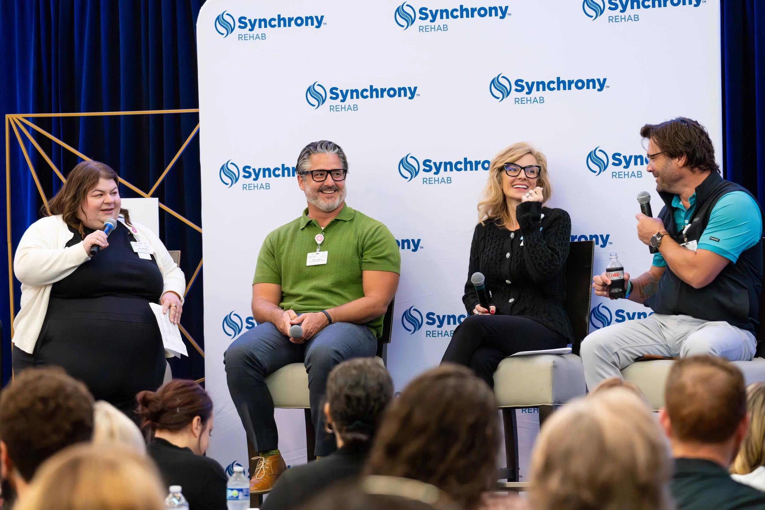 Four people on stage at a panel discussion event with a backdrop featuring the Synchrony Rehab logo, engaging in conversation, smiling, and holding microphones, with an audience in front.