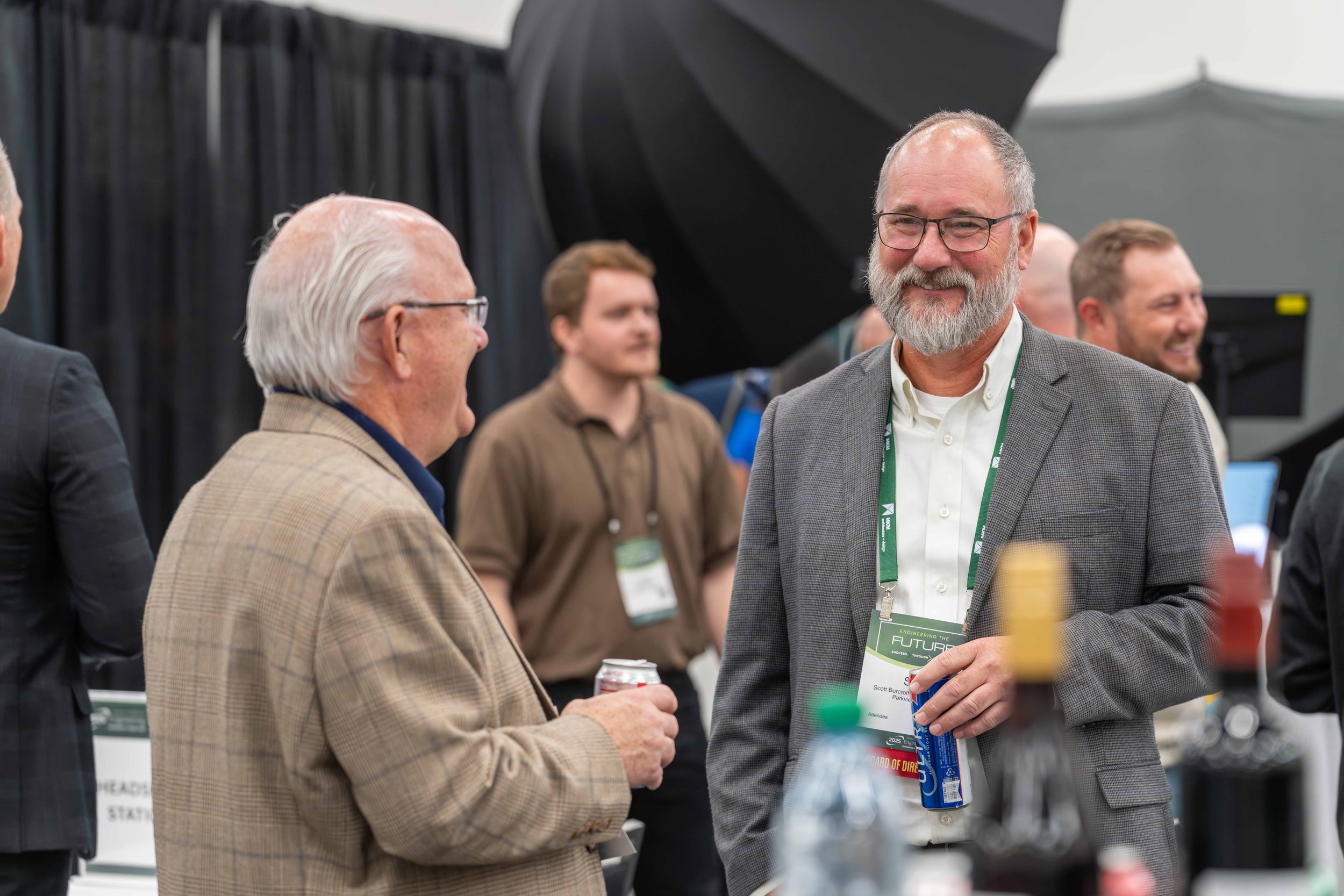 Two men engaged in conversation at a conference, smiling and holding drinks, with other attendees in the background.