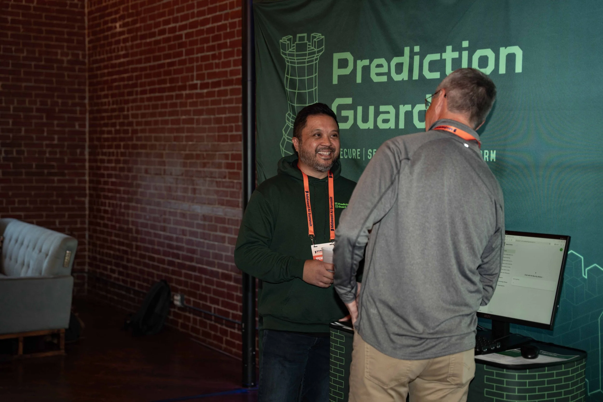 Two men chatting at a booth for Prediction Guard at an event. One man is smiling, holding a cup, and wearing a black hoodie and orange lanyard. The other man has his back to the camera, wearing a gray jacket and glasses. The booth has a backdrop with