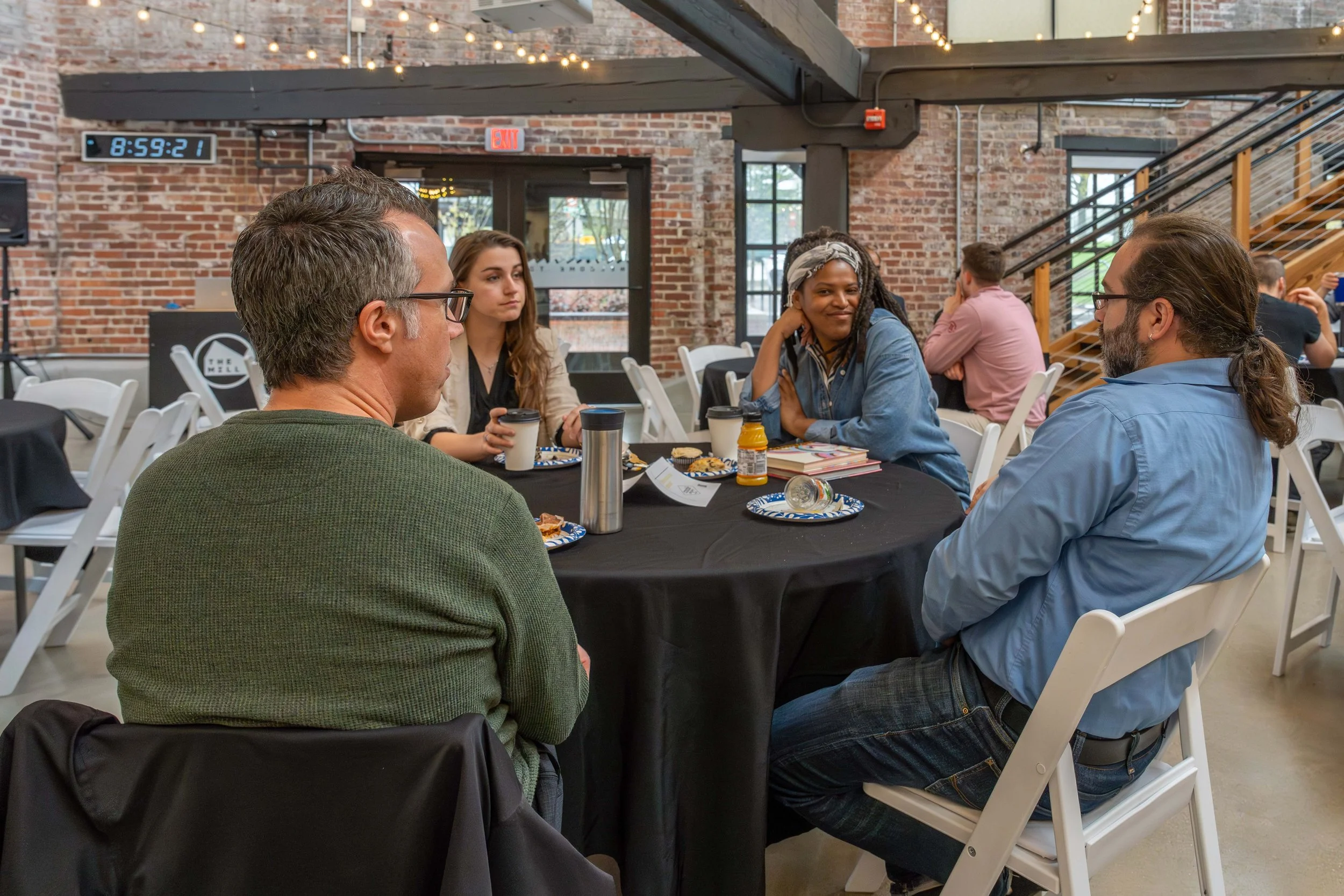 A group of five people seated around a table in a restaurant or cafe, engaged in discussion. There are cups, plates with food, and a bottle on the table. The background features brick walls, large windows, and string lights.