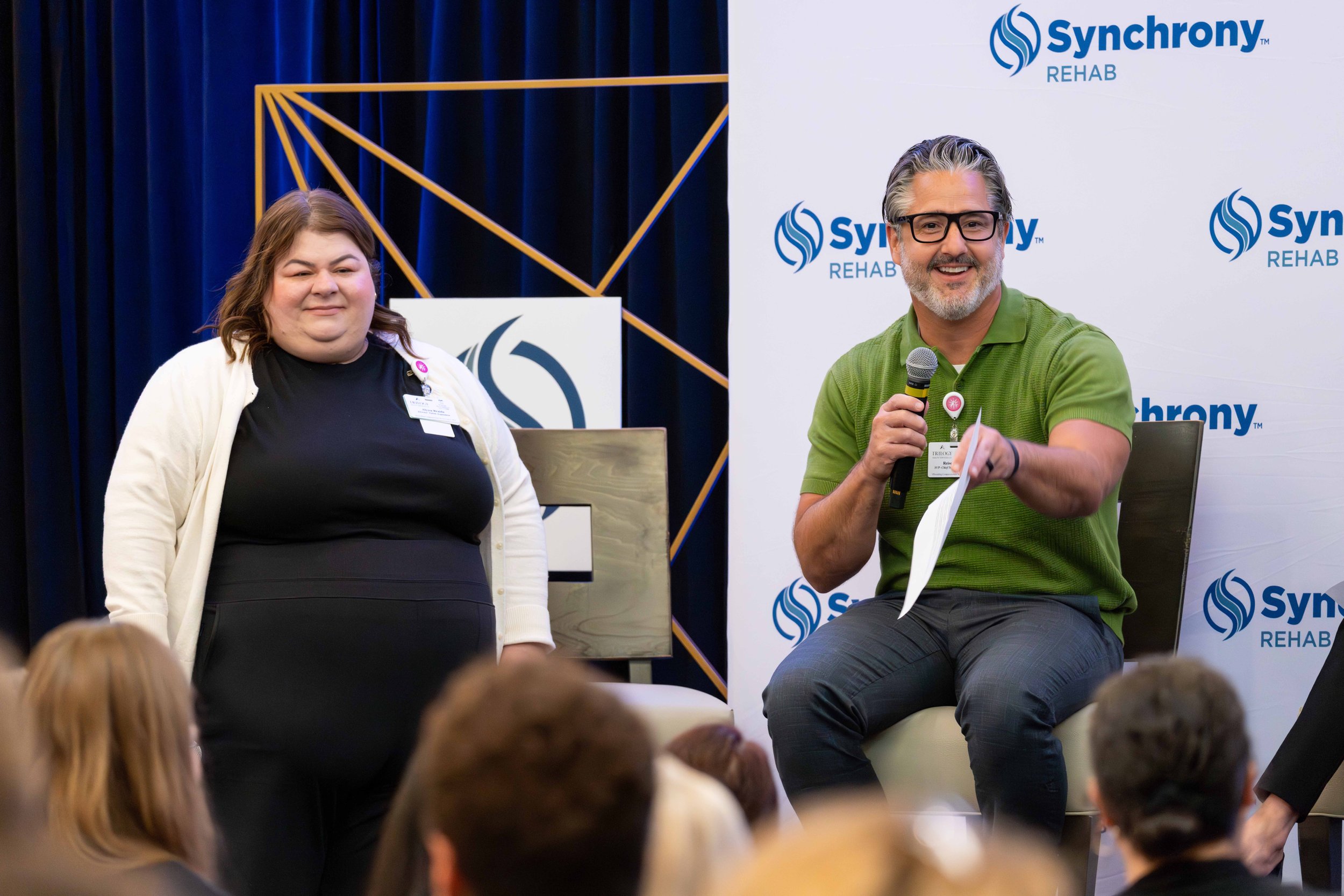 Two people conducting a panel discussion at a conference. A woman on the left stands while a man on the right sits with a microphone, both smiling. Background displays the logos of Synchronic Rehab.